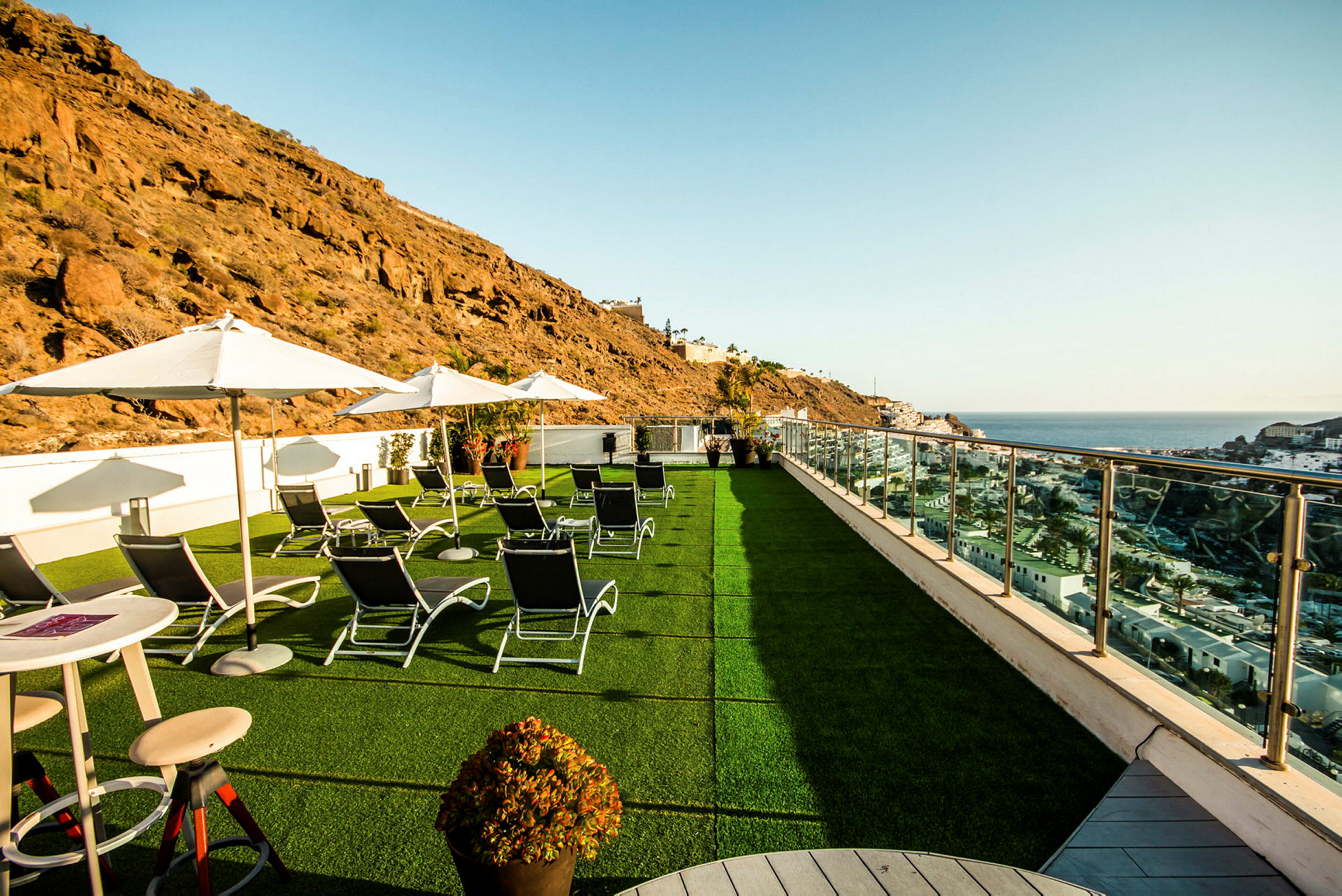 Billede af en terrasse med liggestole, parasoller og udsigt over havet og omkringliggende klipper. (AI-genereret tekst)