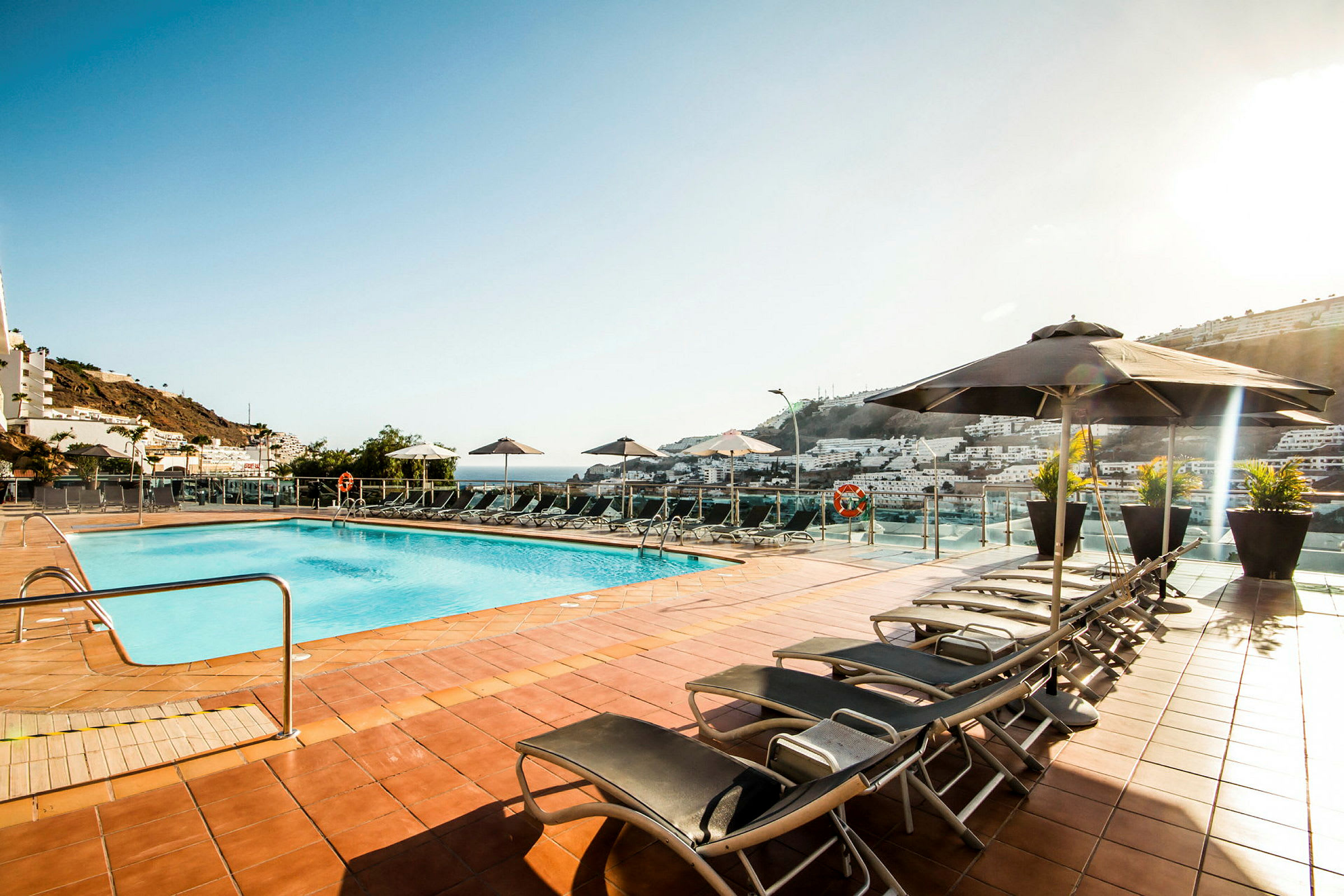 En udendørs swimmingpool med liggestole og parasoller rundt om, beliggende på en terrasse med udsigt over en by og naturskønne omgivelser. (AI-genereret tekst)
