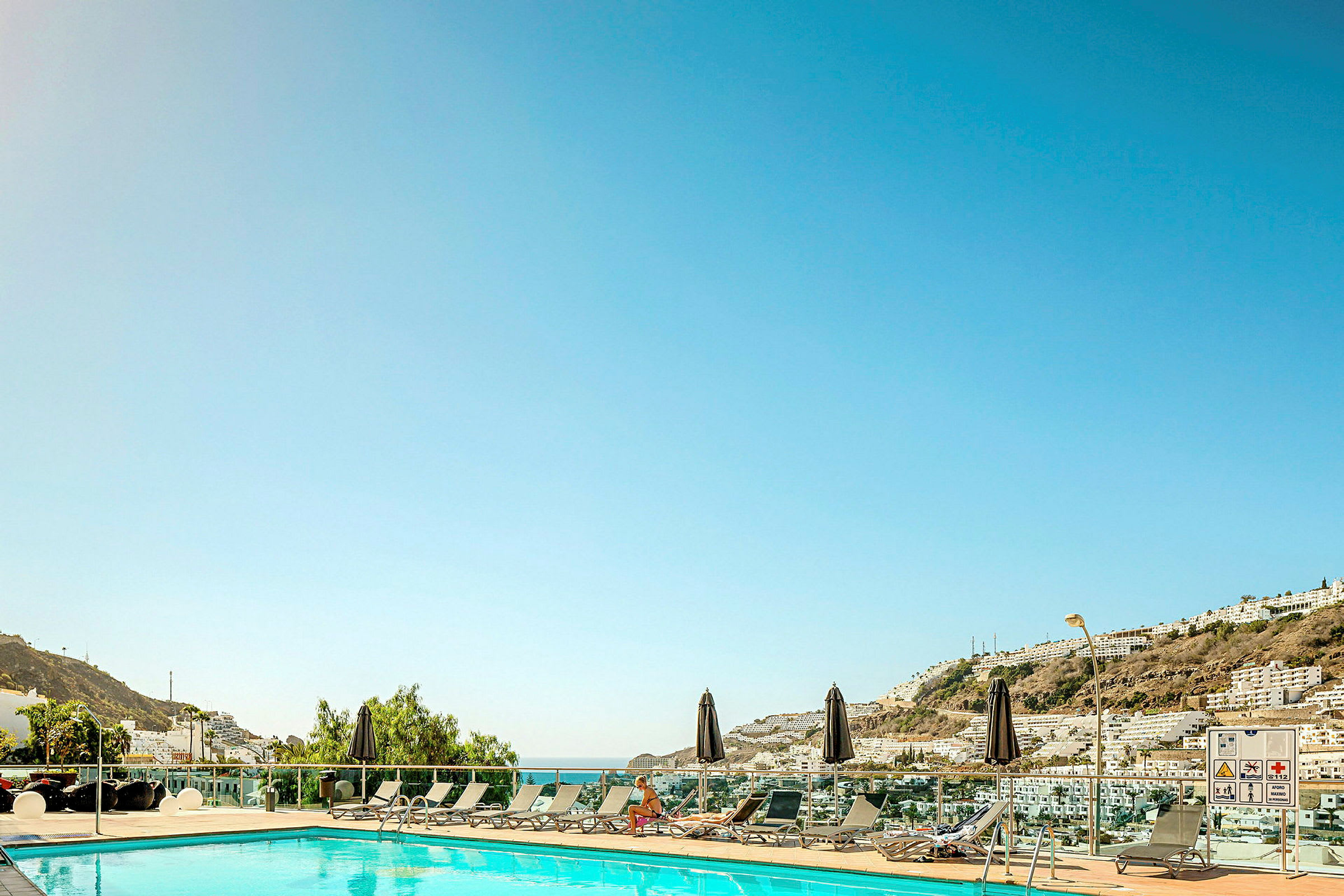 En udendørs pool omgivet af liggestole og parasoller, med udsigt over en by og bjerge i baggrunden under en klar himmel. (AI-genereret tekst)