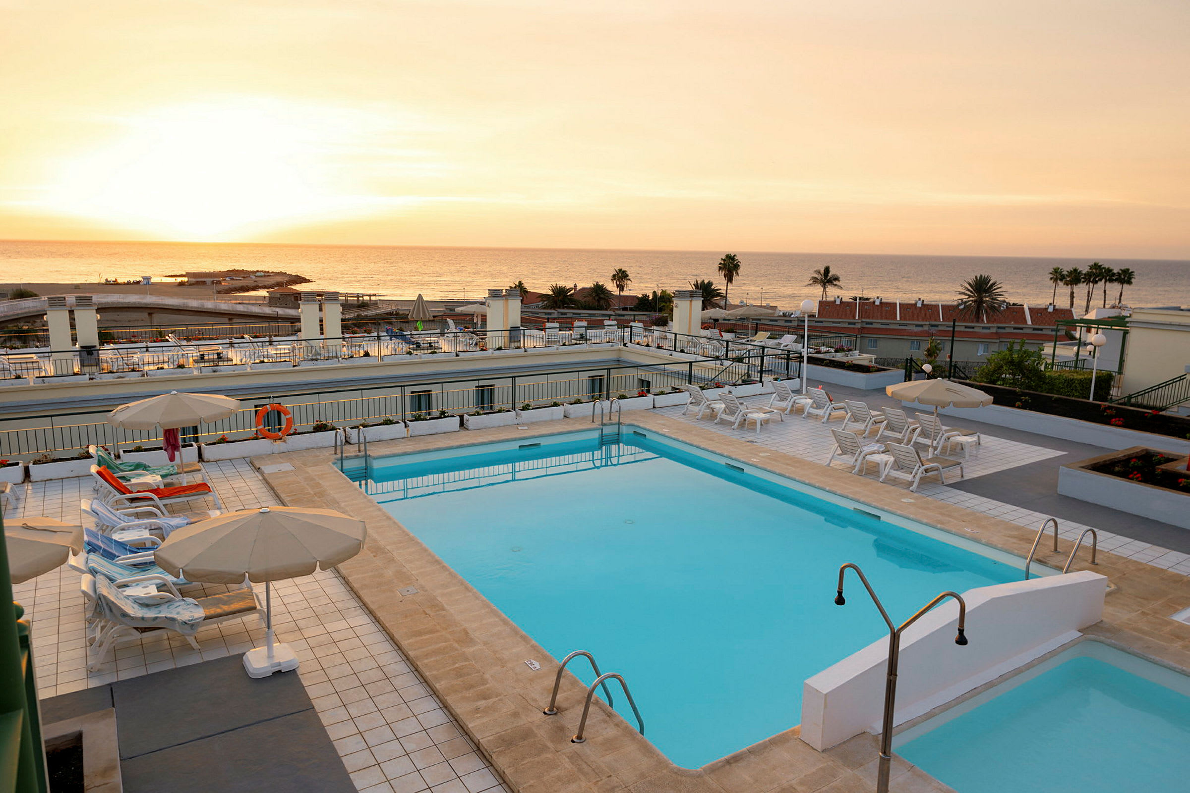 En udendørs swimmingpool med liggestole og parasoller ved solnedgang, med havet i baggrunden. (AI-genereret tekst)