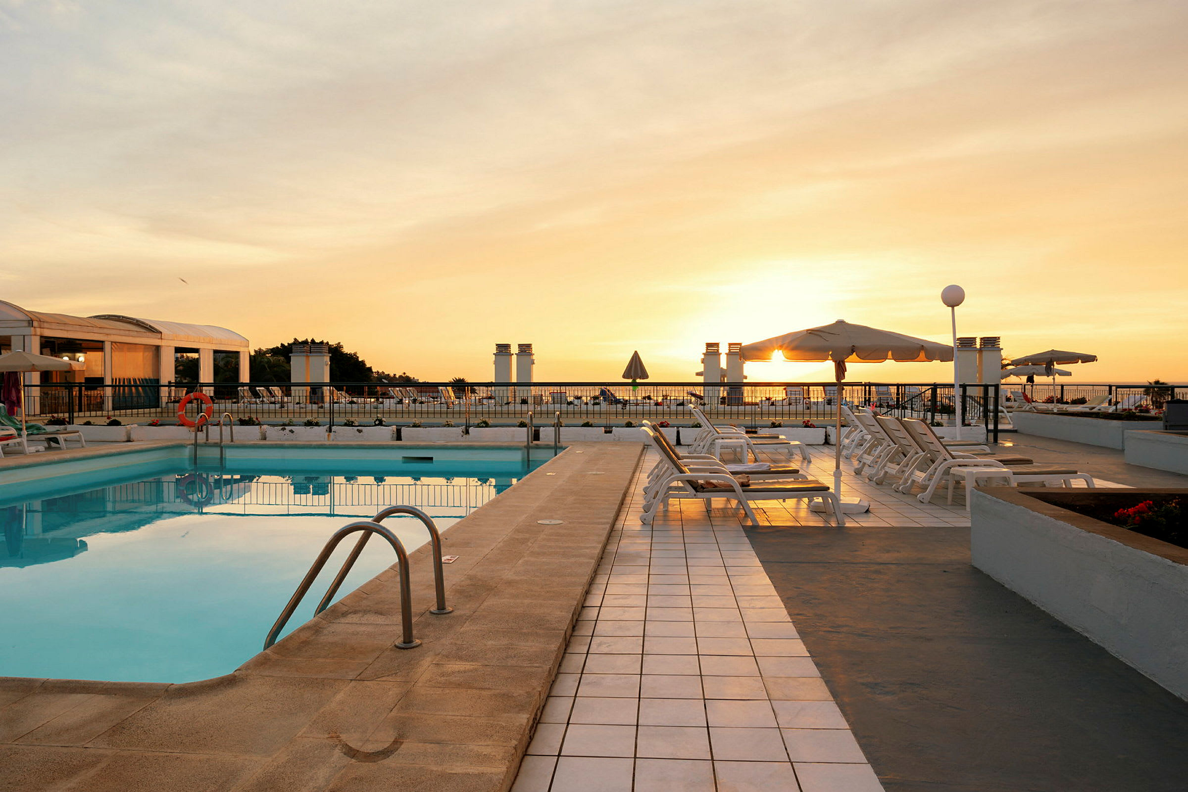 Udendørs poolområde i solnedgang med liggestole og parasoller. (AI-genereret tekst)