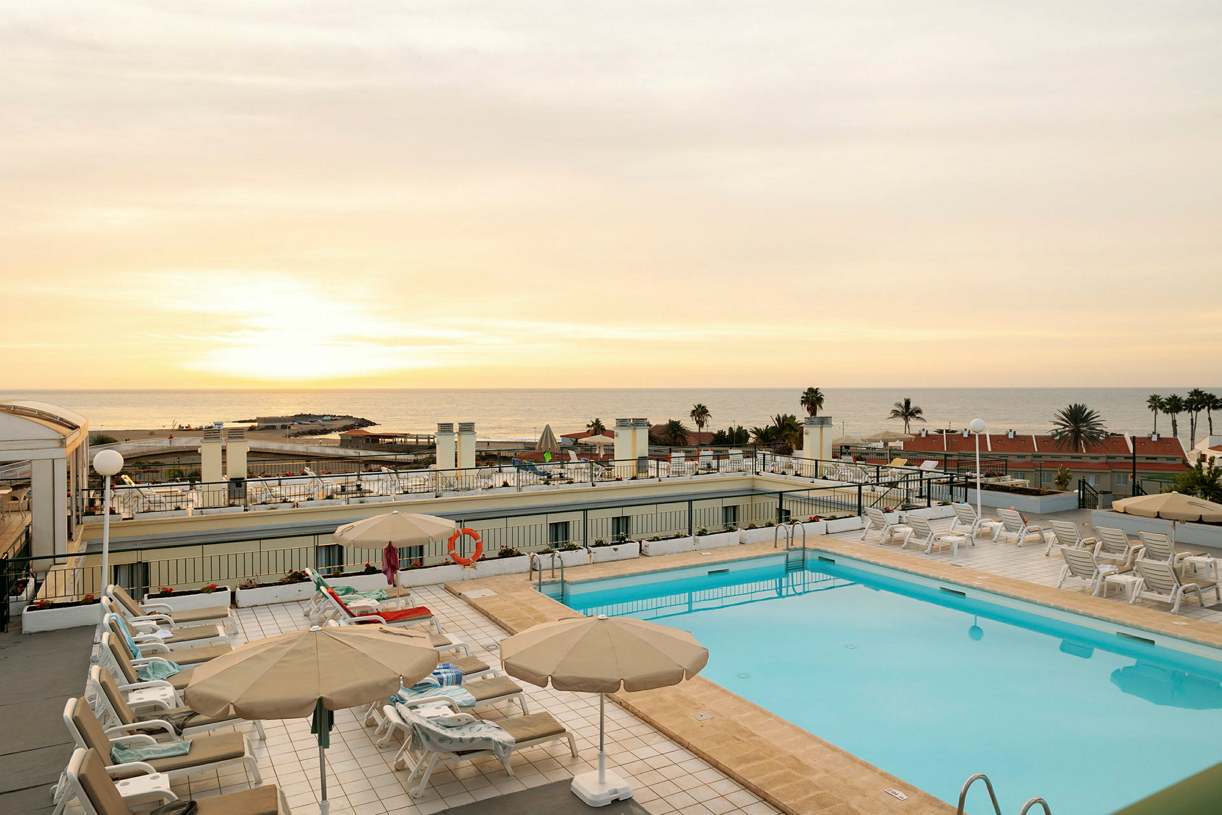 En hotelpool med liggestole og parasoller med udsigt over havet ved solnedgang. (AI-genereret tekst)