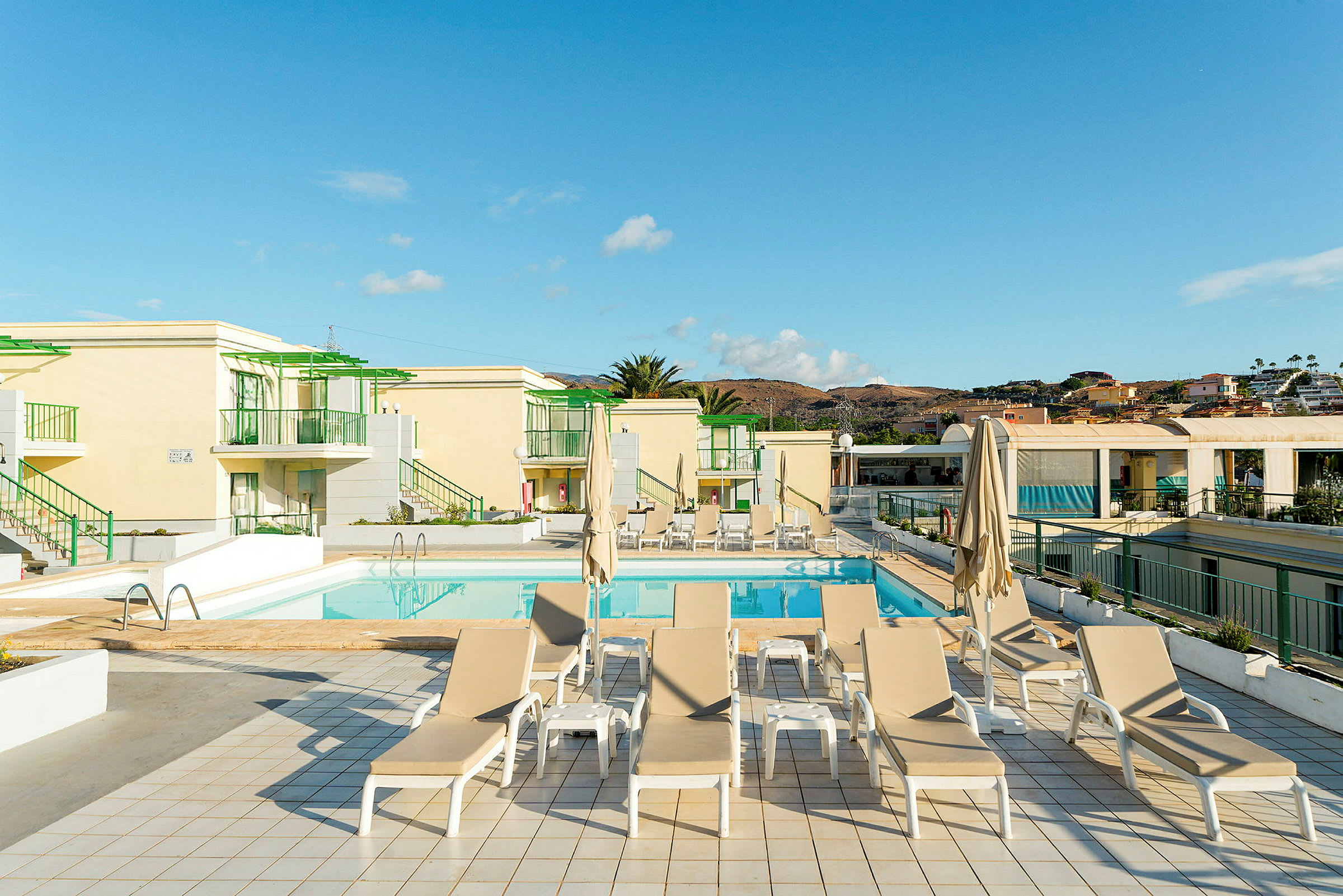 En solterrasse ved en pool med liggestole og parasoller, omgivet af bygninger. (AI-genereret tekst)
