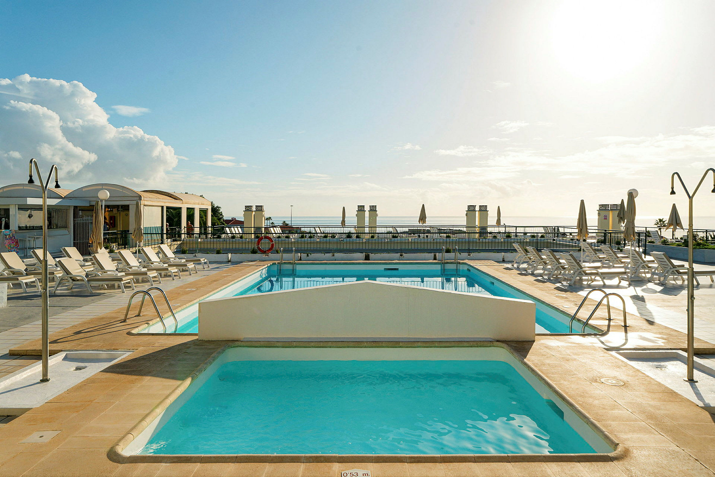 En udendørs pool med liggestole og parasoller under en solrig himmel. (AI-genereret tekst)