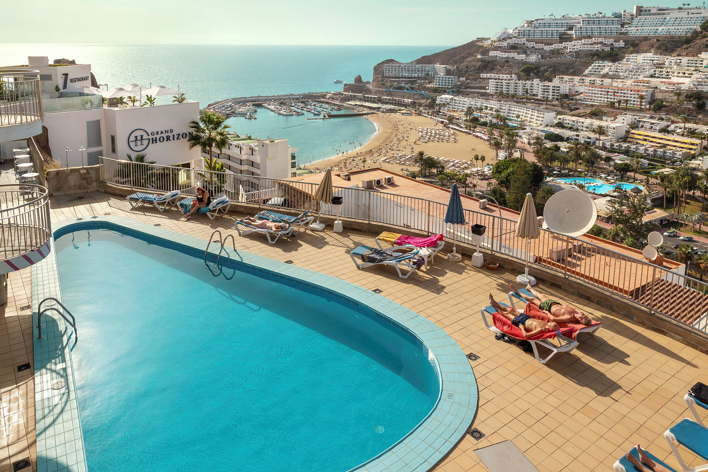 En udendørs pool med liggestole på en terrasse, med udsigt over en strand og havet. (AI-genereret tekst)