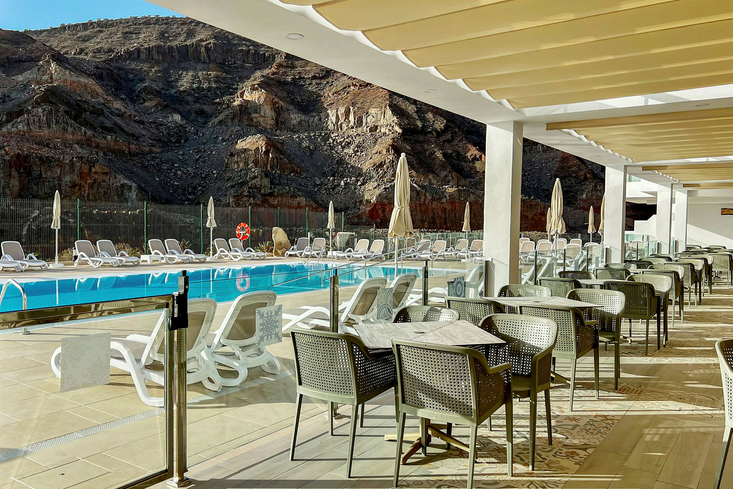 Udsigt over en pool med solsenge og parasoller foran en bjergagtig baggrund samt spiseområde med borde og stole på en terrasse. (AI-genereret tekst)