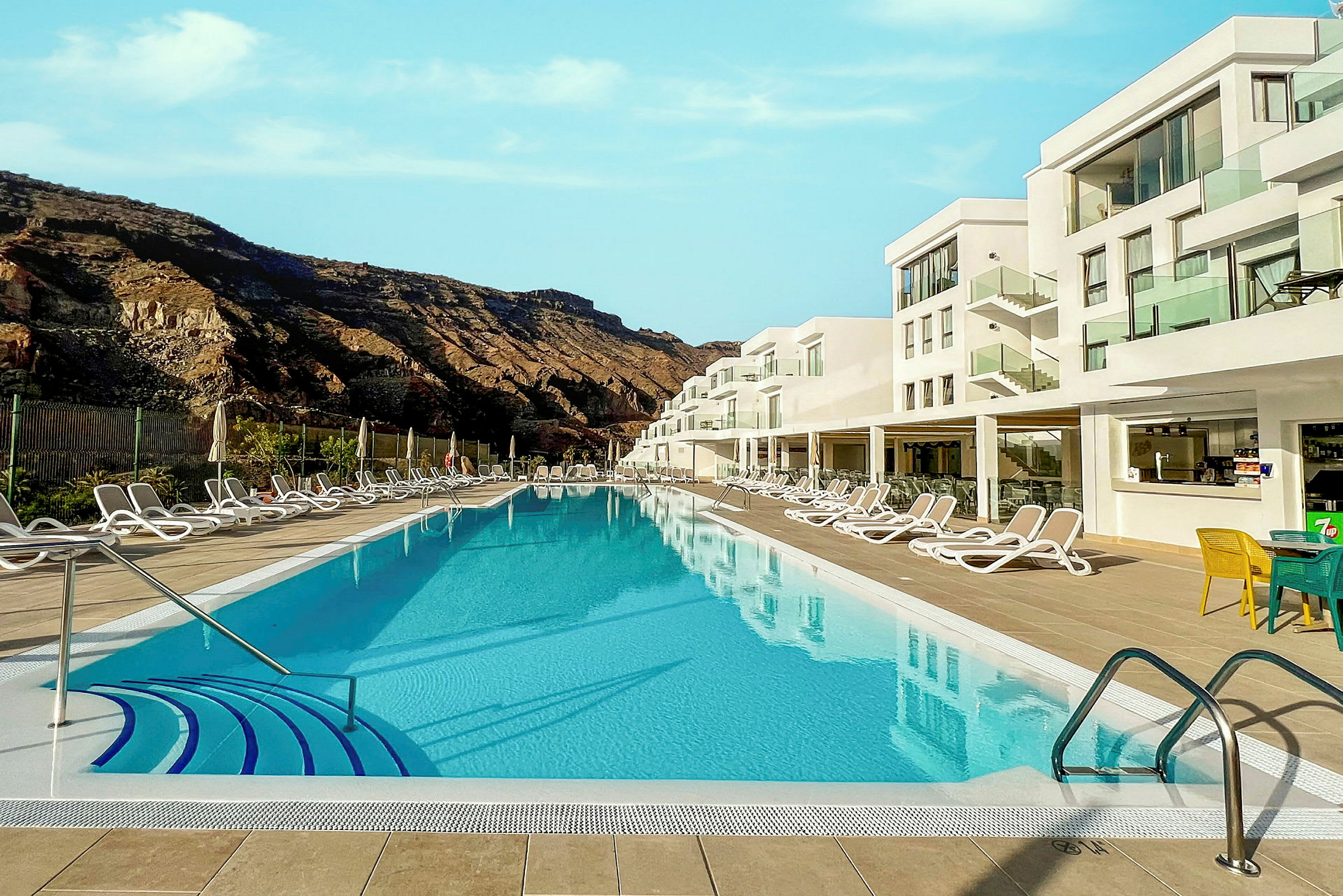 En pool omgivet af liggestole og en moderne hotelbygning i baggrunden. (AI-genereret tekst)