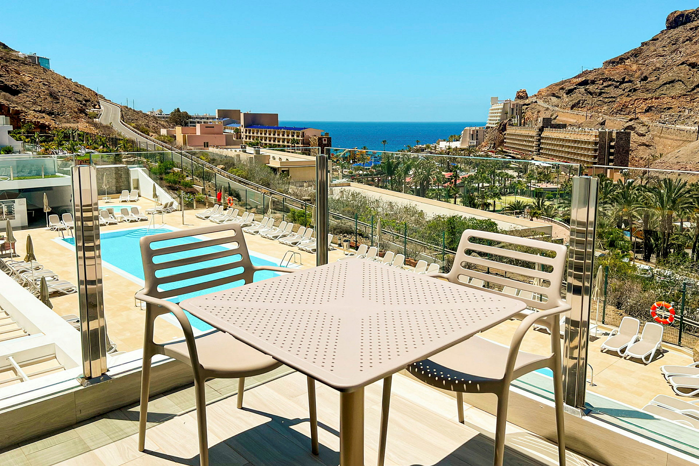 Udsigt over en pool og solsenge med havet og bygninger i baggrunden, set fra en balkon med bord og stole. (AI-genereret tekst)