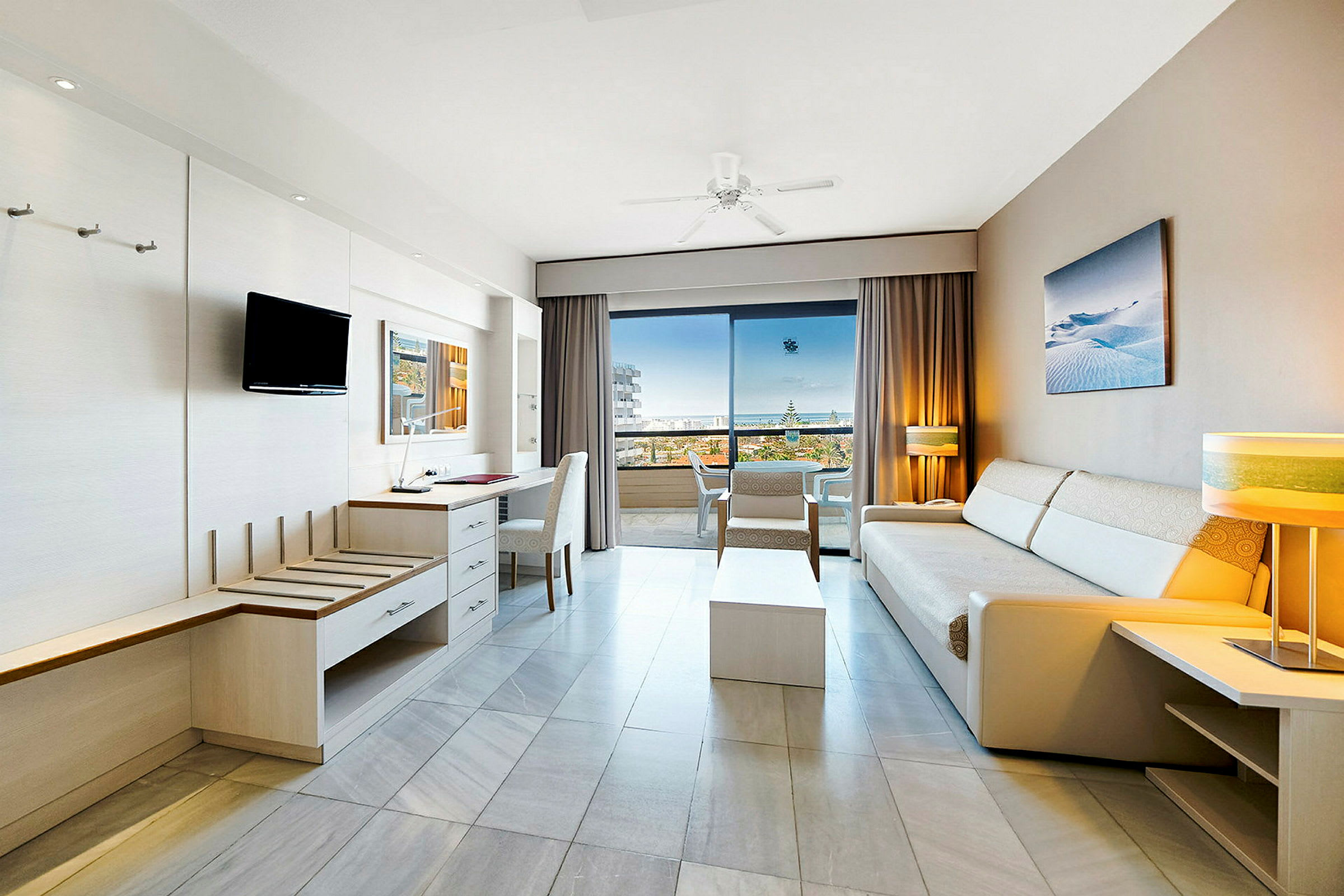 Lyst hotelværelse med sofaer og møbler, åbent mod en balkon med havudsigt. (AI-genereret tekst)