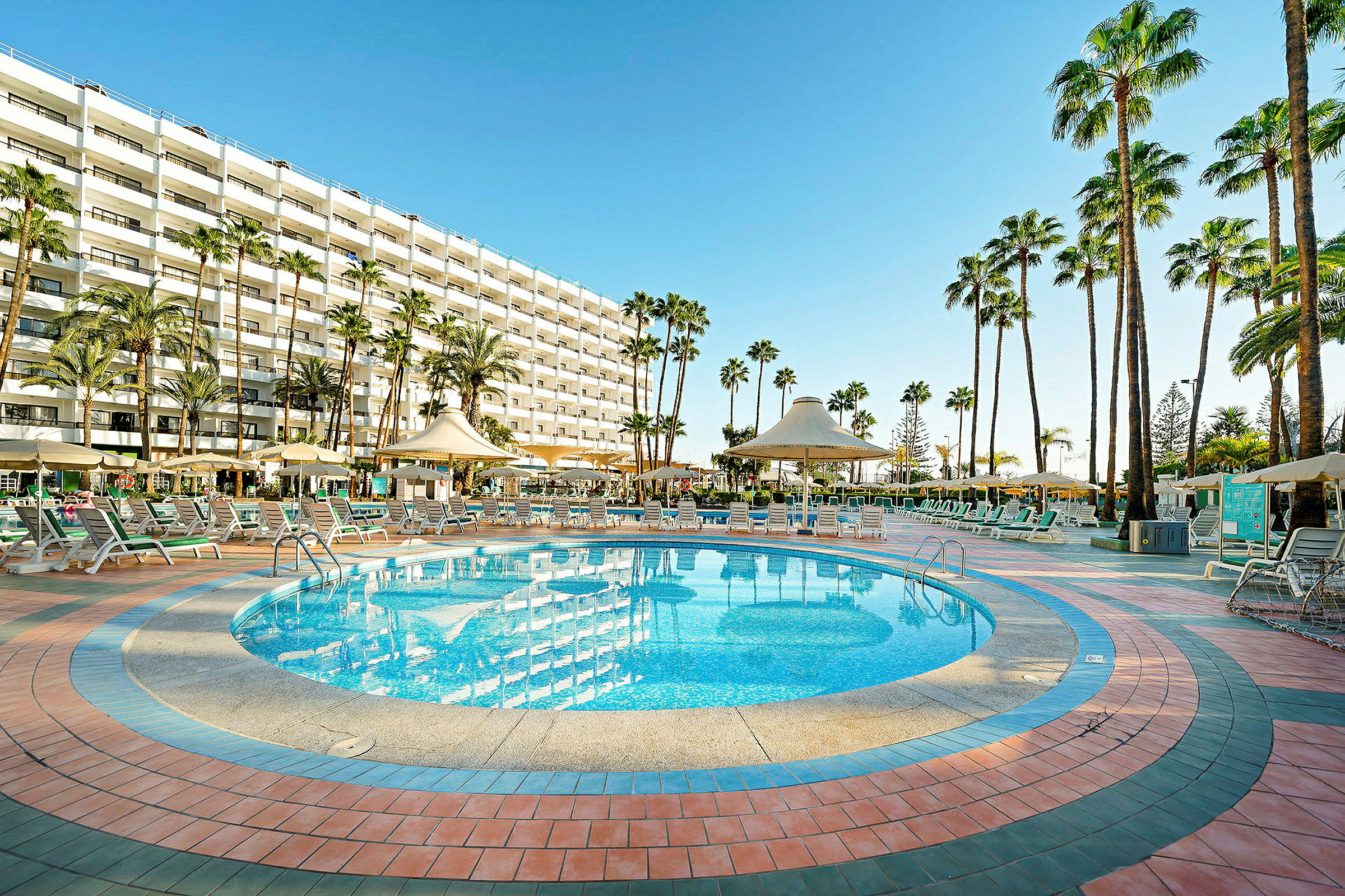 Udendørs swimmingpool med liggestole og parasoller, omgivet af palmer og en hotelbygning i baggrunden. (AI-genereret tekst)