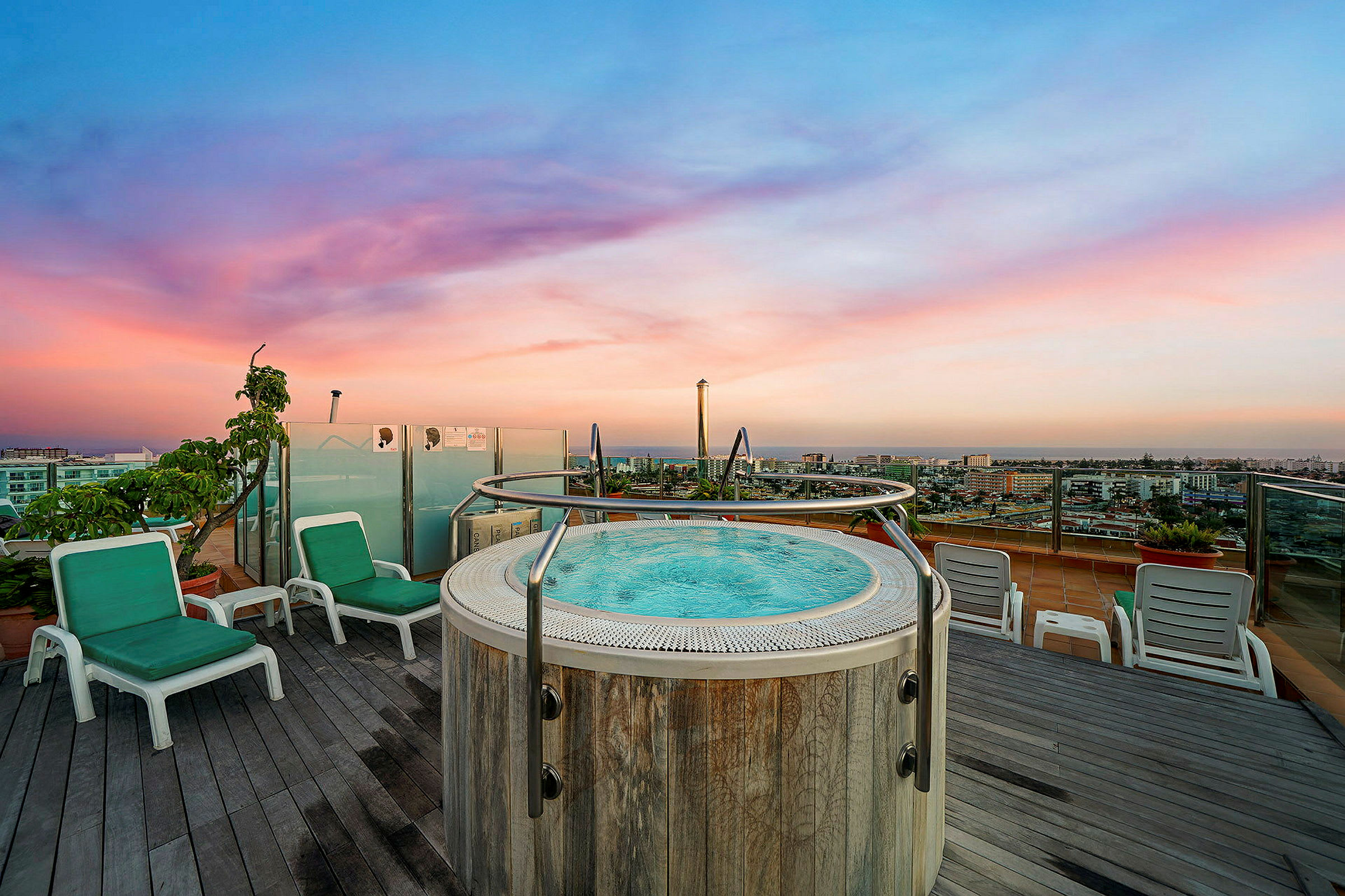 Billedet viser et boblebad på en hotelterrasse med udsigt over en by ved solnedgang. (AI-genereret tekst)
