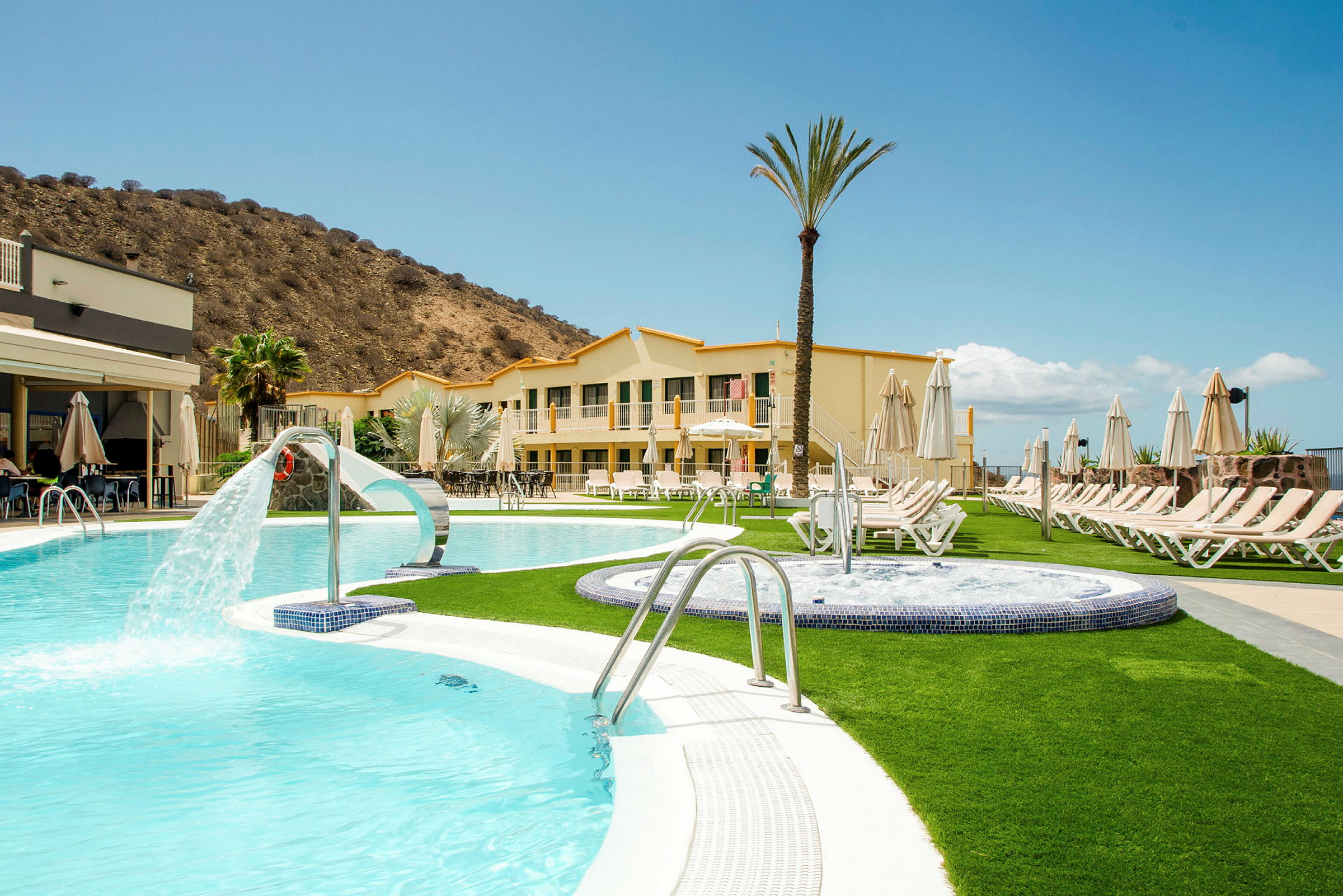 Et hotel med swimmingpools og solsenge, beliggende tæt på et bjerg. (AI-genereret tekst)