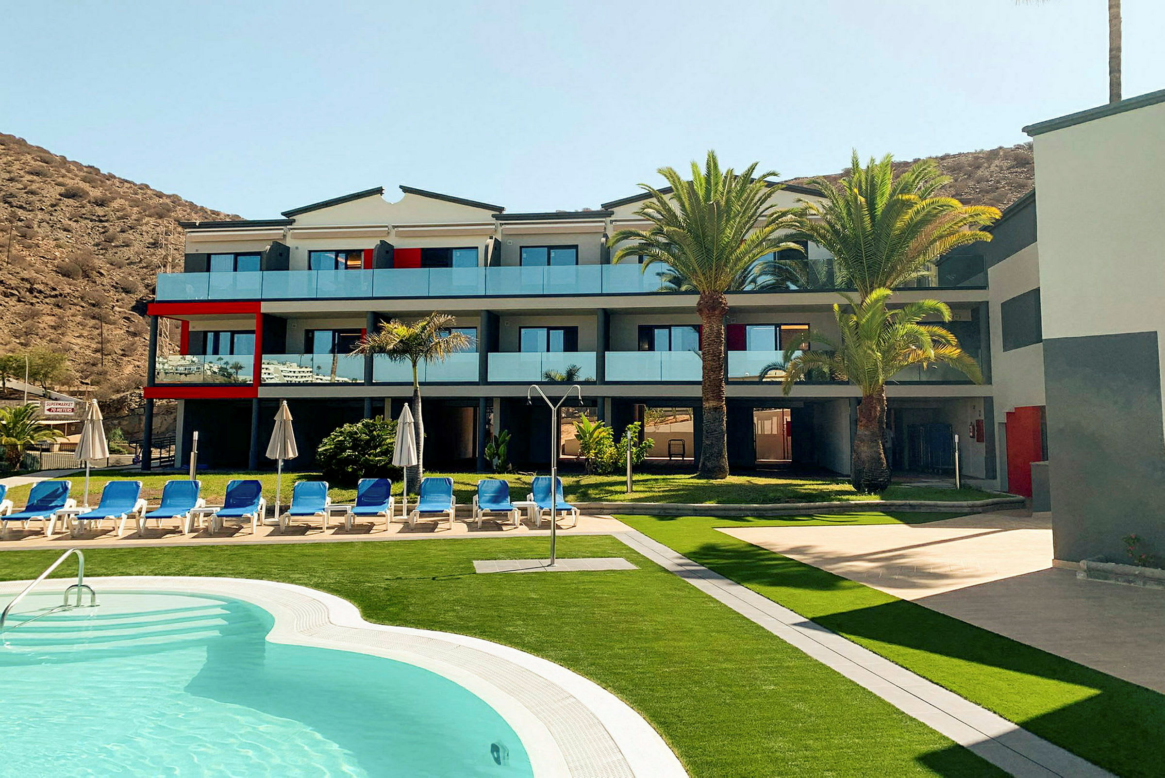 Moderne hotel med pool og liggestole, omgivet af palmer. (AI-genereret tekst)