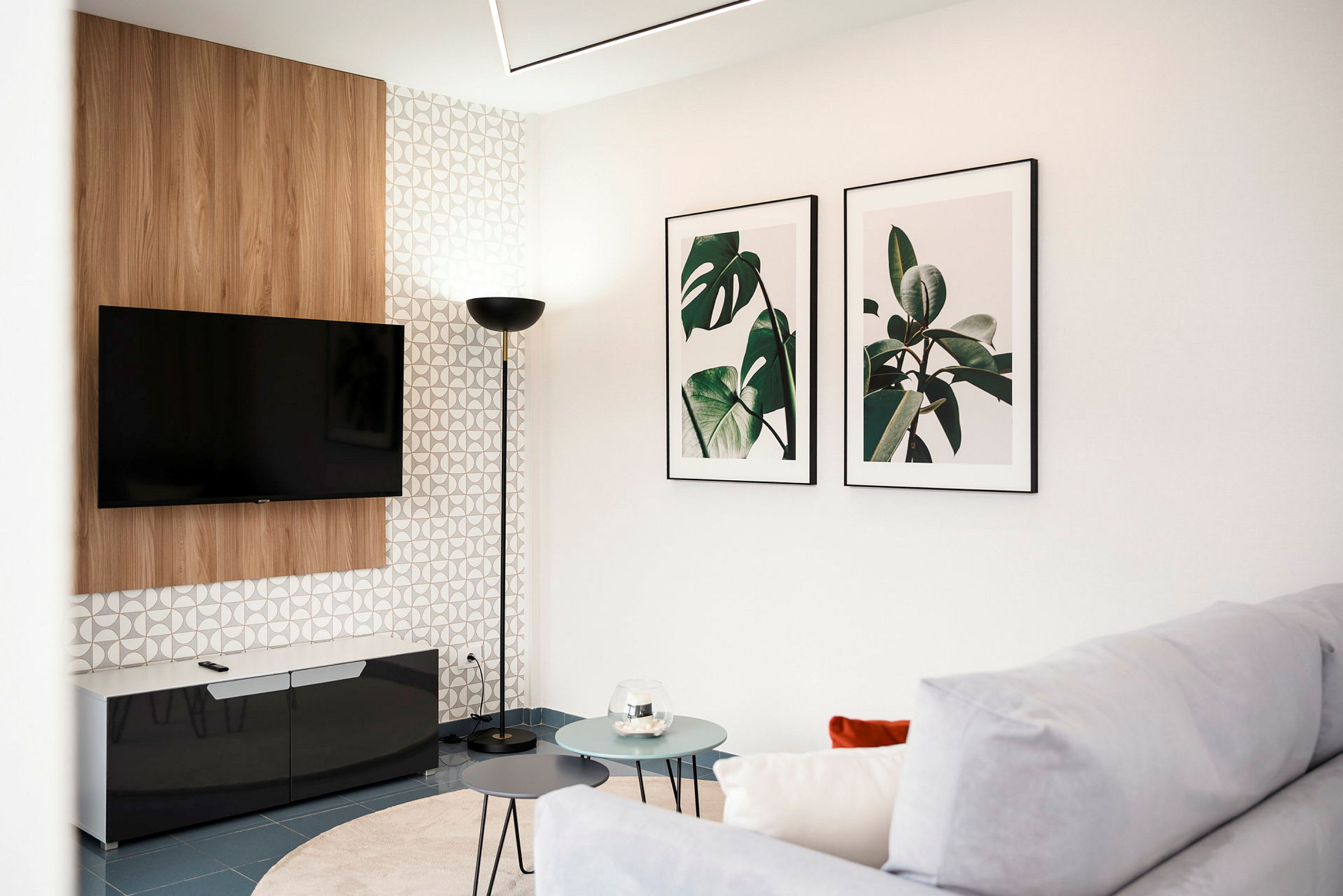En moderne stue med et vægmonteret TV, to billeder med plantemotiver på væggen og et hjørnebord med runde bordplader foran en sofa. (AI-genereret tekst)
