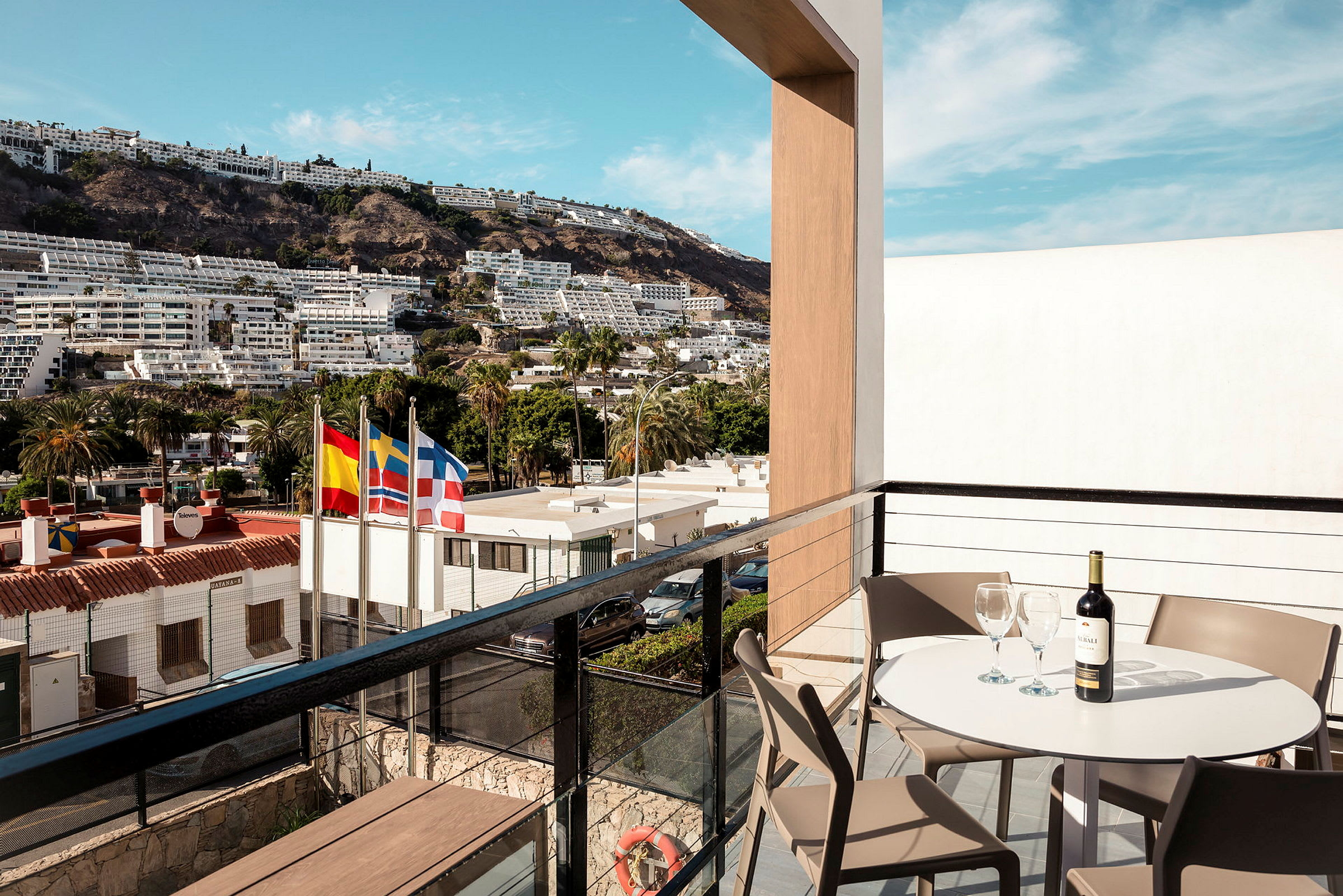 Udsigt fra en balkon med bord, stole og en flaske vin, med flag og bygninger i baggrunden. (AI-genereret tekst)