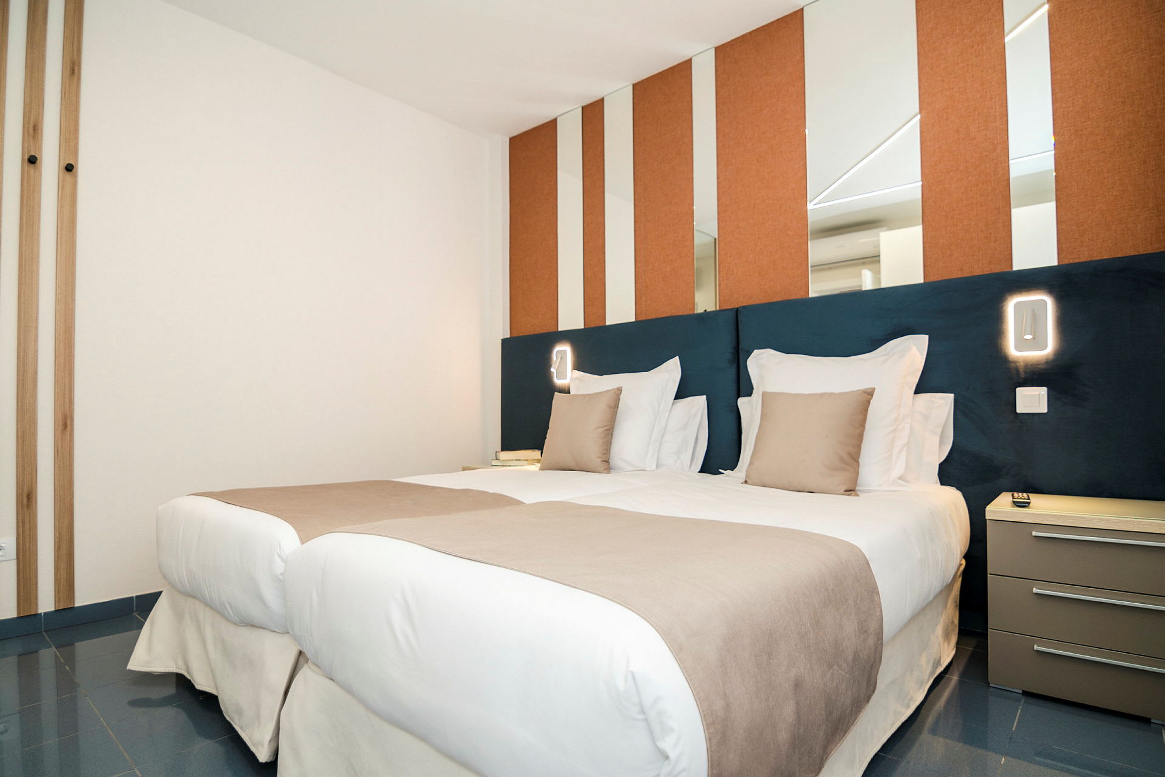 Et moderne hotelværelse med to enkeltsenge side om side, redt med hvide og beige sengetøj. Væggene har spejle og orange paneler, og ved siden af sengen er et natbord. (AI-genereret tekst)