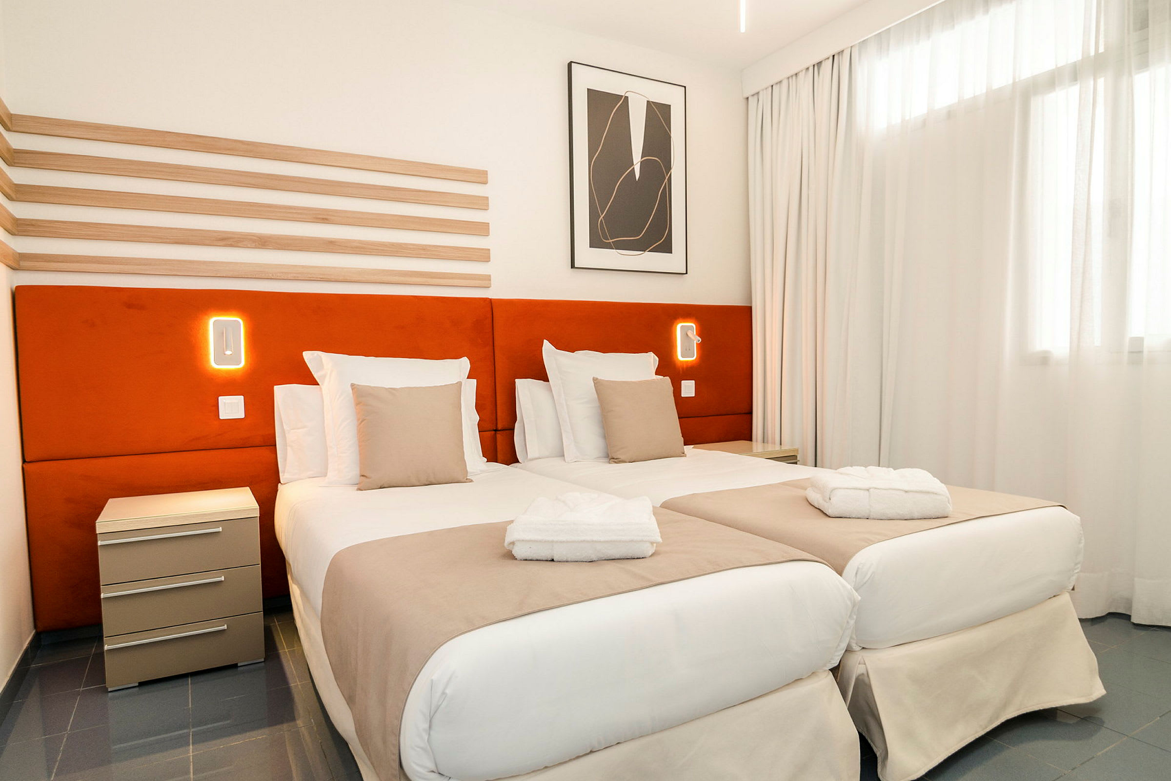 Et hotelværelse med to enkeltsenge, orange sengegærder og hvidt sengetøj. En natlampe ses på væggen. (AI-genereret tekst)
