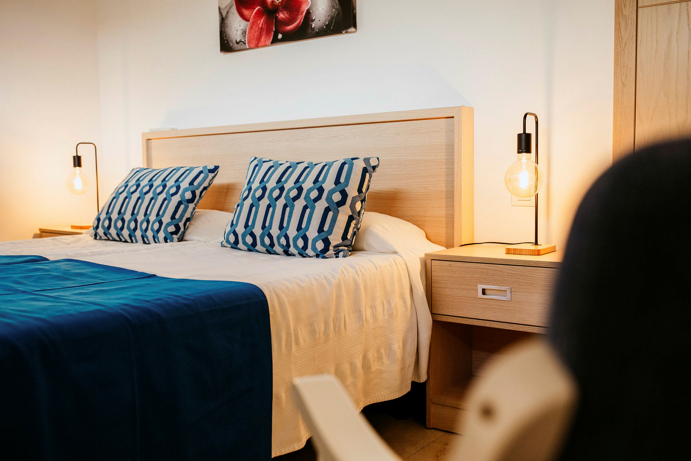 Dobbeltværelse med blå puder og sengetæppe, natbord med en lampe. (AI-genereret tekst)