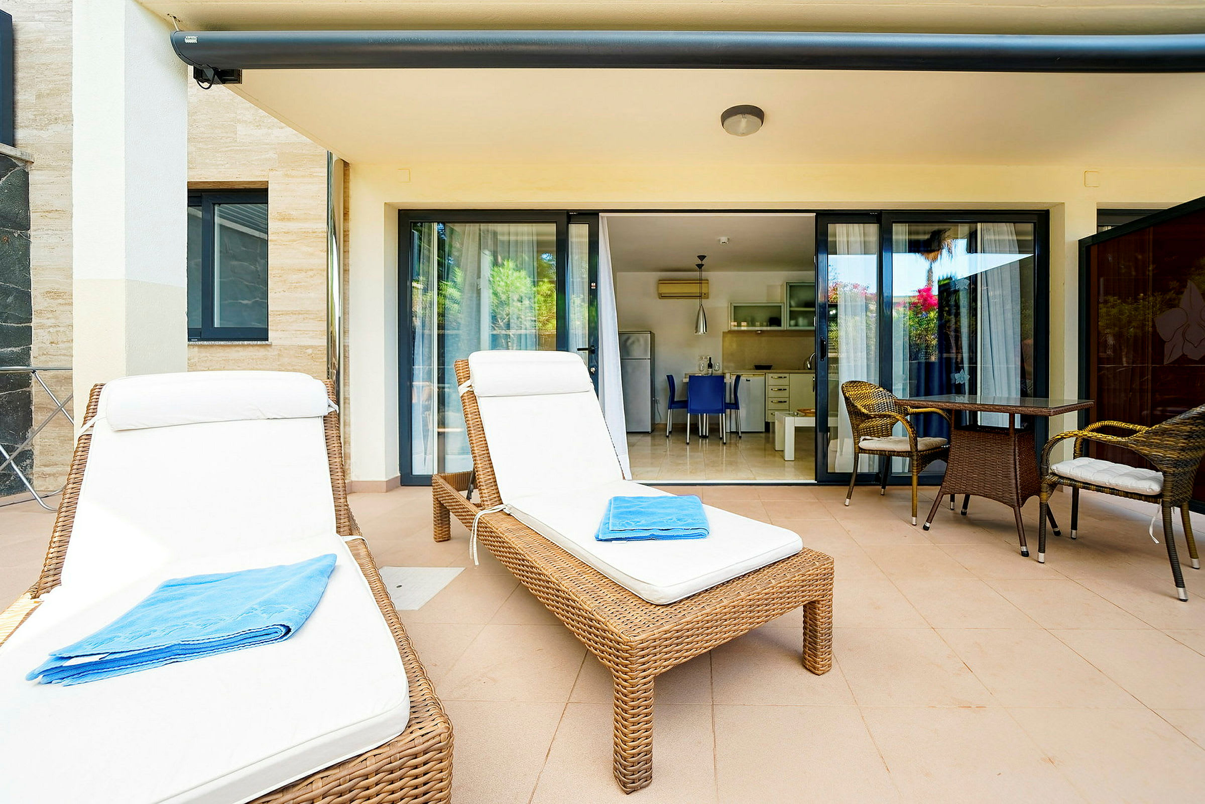 To liggestole med hvide hynder og et blåt håndklæde på en terrasse med bord og stole foran glasdøre til et køkken. (AI-genereret tekst)