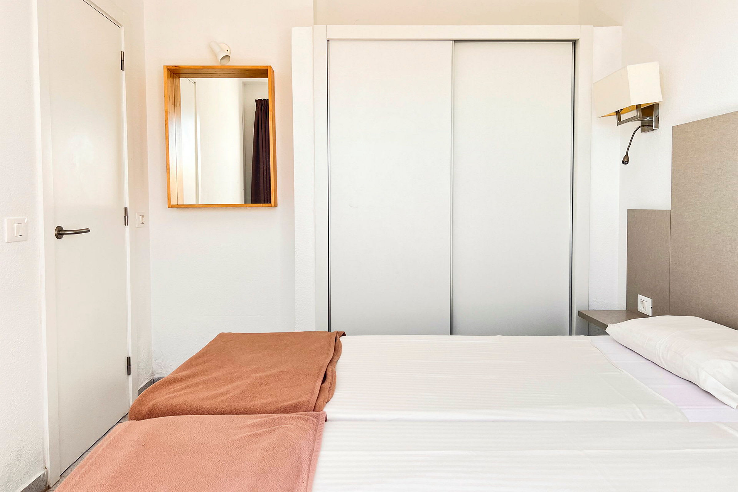Et hotelværelse med hvidt interiør, en dobbeltseng med hvide lagner og brune sengetæpper samt et spejl på væggen. (AI-genereret tekst)