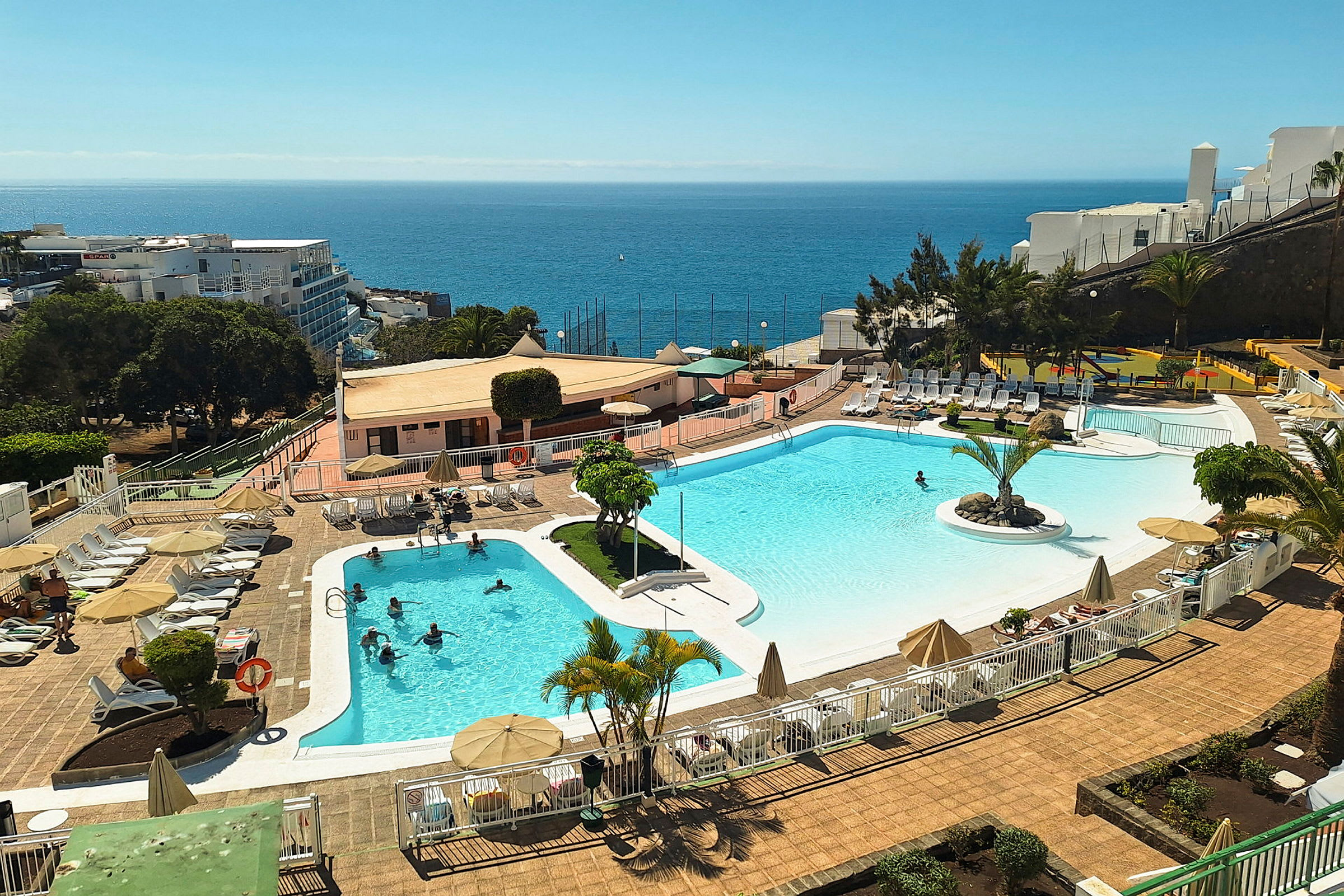 En udendørs pool ved et feriehotel med liggestole og parasoller. Havet ses i baggrunden. (AI-genereret tekst)