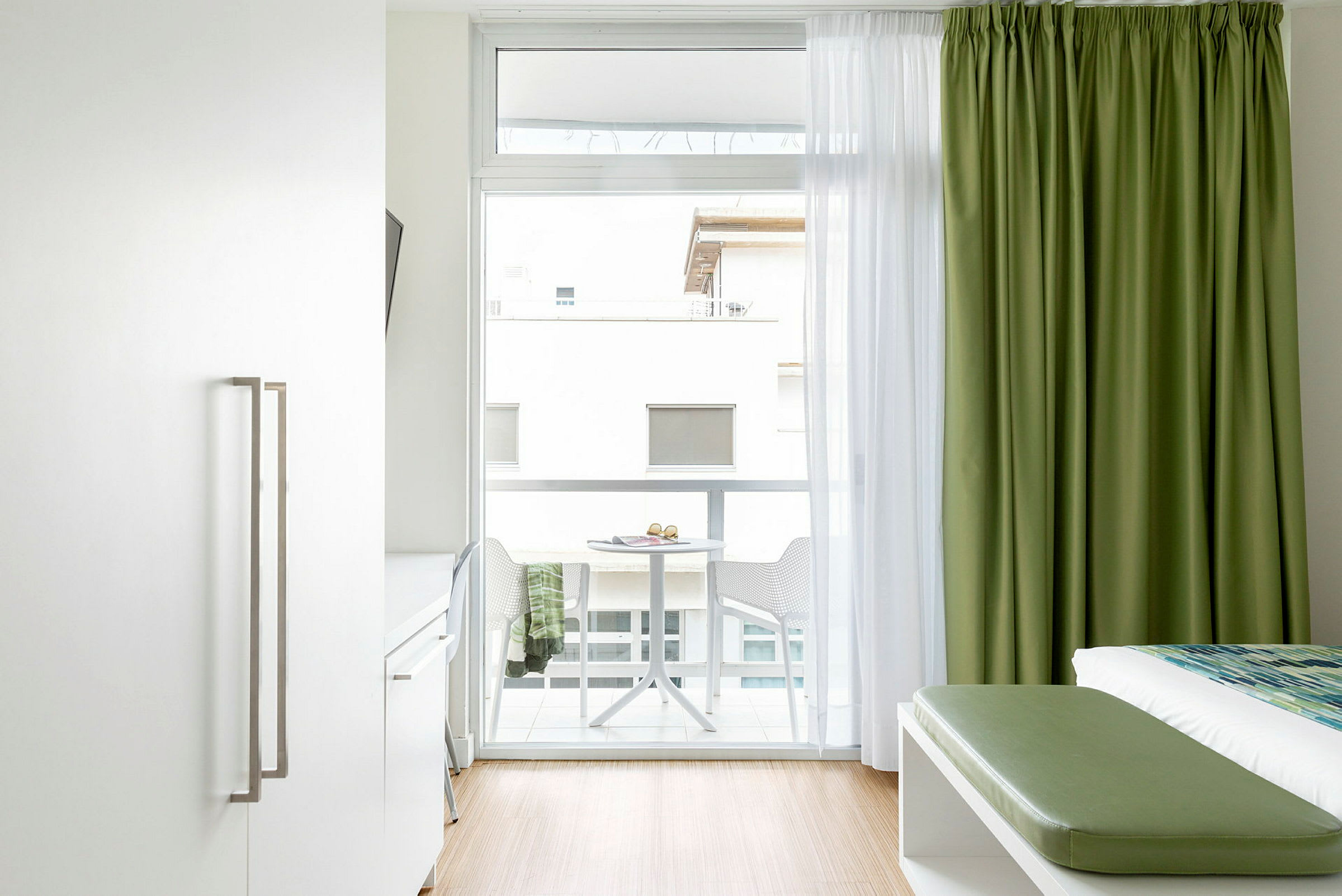 Et lyst hotelværelseinteriør med en seng, grønne gardiner og en altan med bord og stole. (AI-genereret tekst)