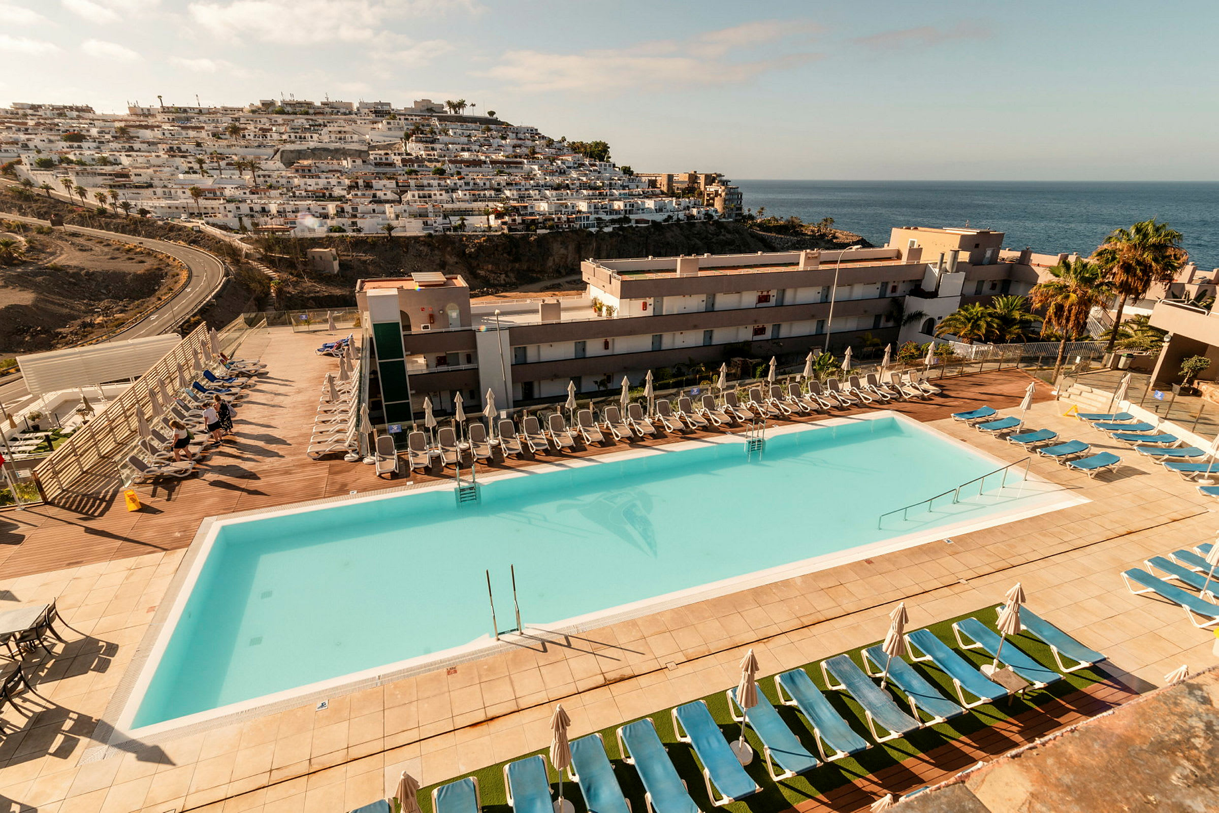 En udendørs pool med solsenge og parasoller ved et hotelområde, med havudsigt og en bjergskråning med bygninger. (AI-genereret tekst)