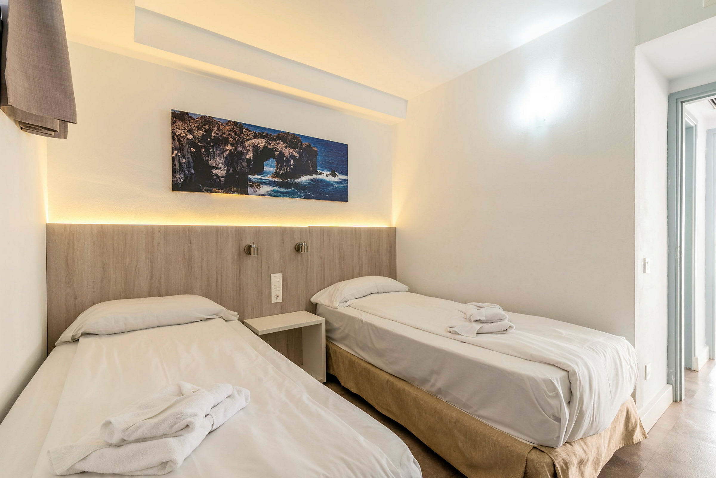To enkeltsenge med hvide lagner og håndklæder på et lyst hotelværelse med et klippemaleri ovenover. (AI-genereret tekst)