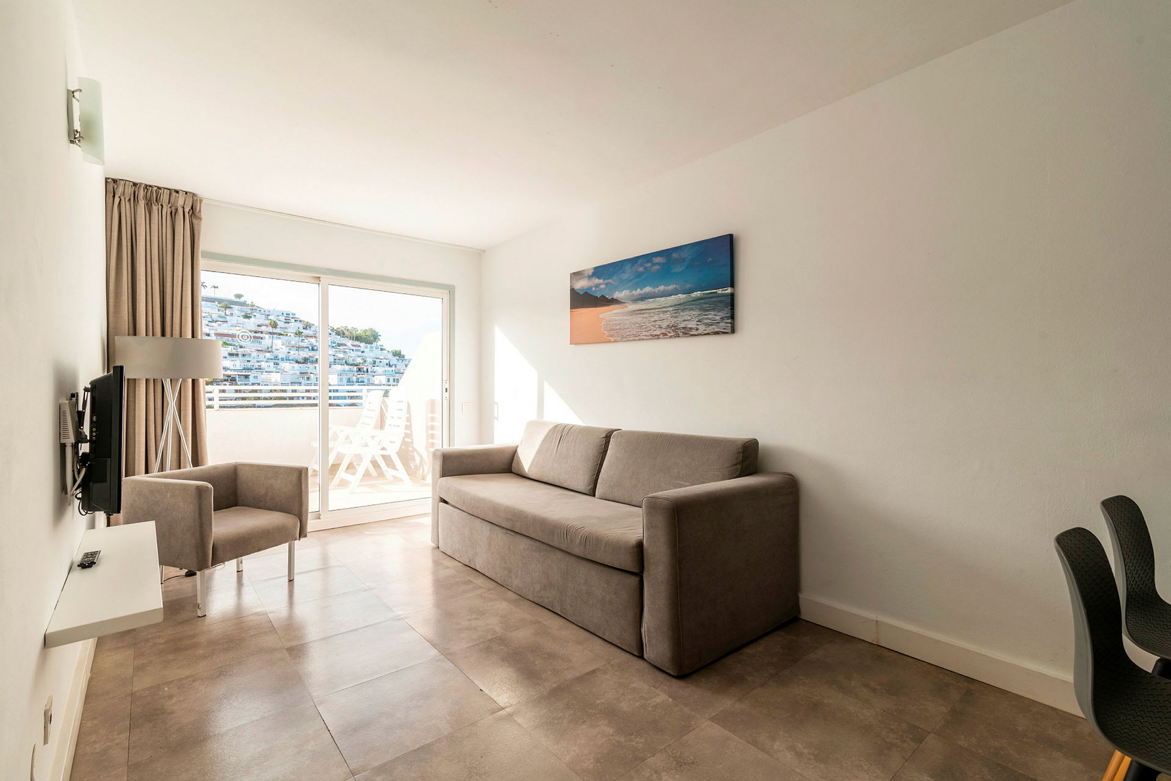 Et hotelværelse med en beige sofa og en lænestol samt en altan med udsigt. (AI-genereret tekst)