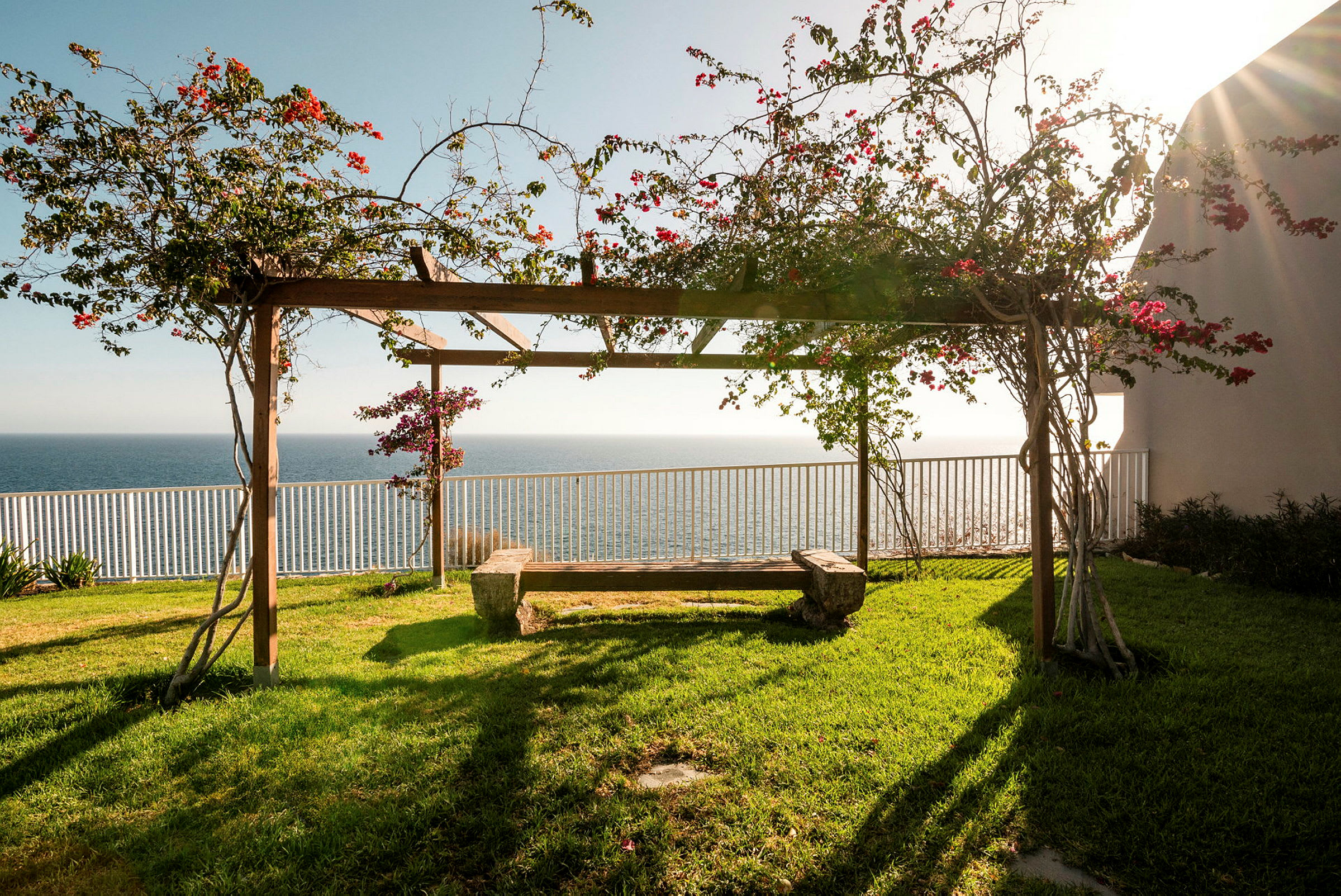 En pergola med blomstrende planter og bænk, havet i baggrunden. (AI-genereret tekst)