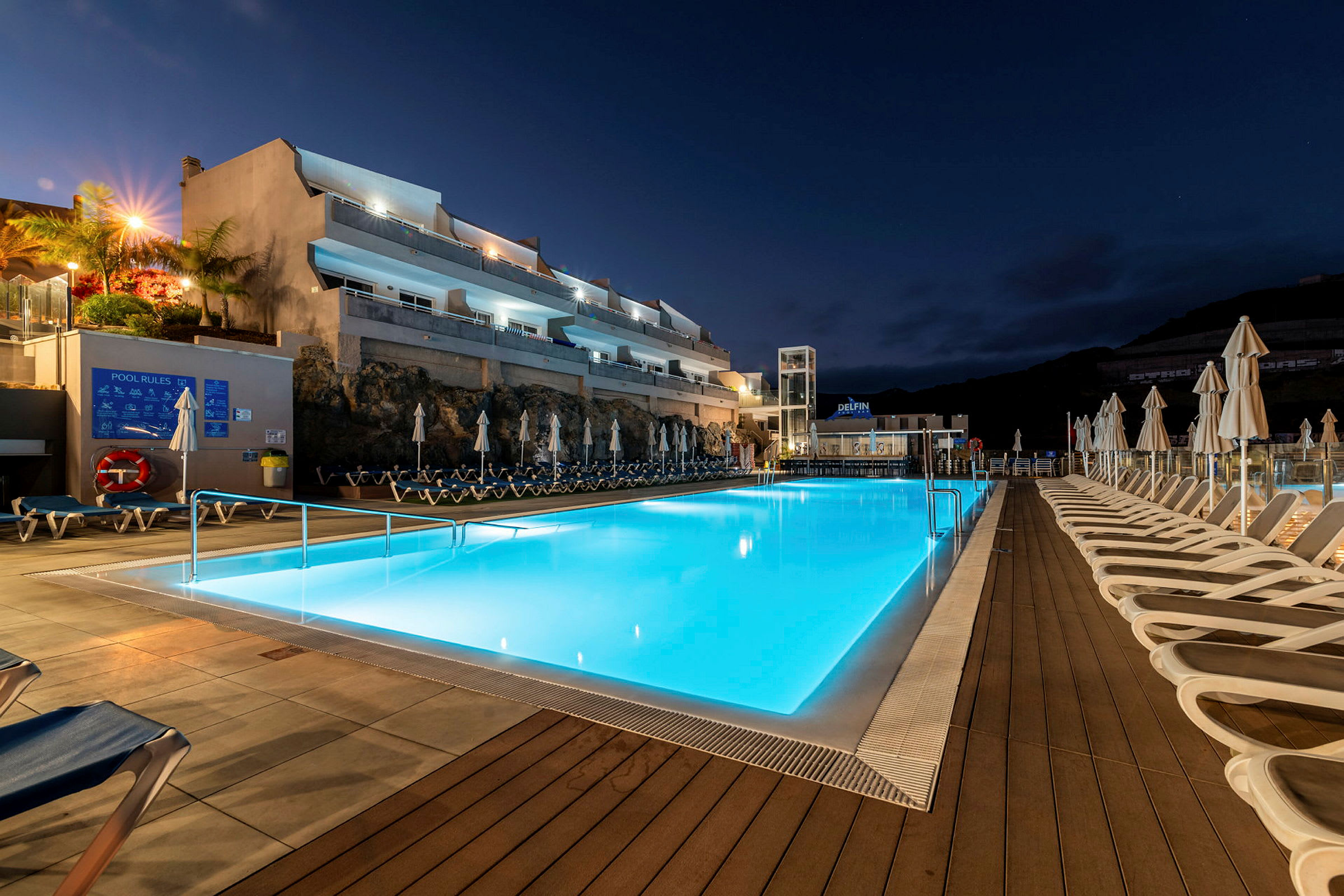Udendørs udsigt over en oplyst swimmingpool om natten, omgivet af liggestole og bygninger i baggrunden. (AI-genereret tekst)