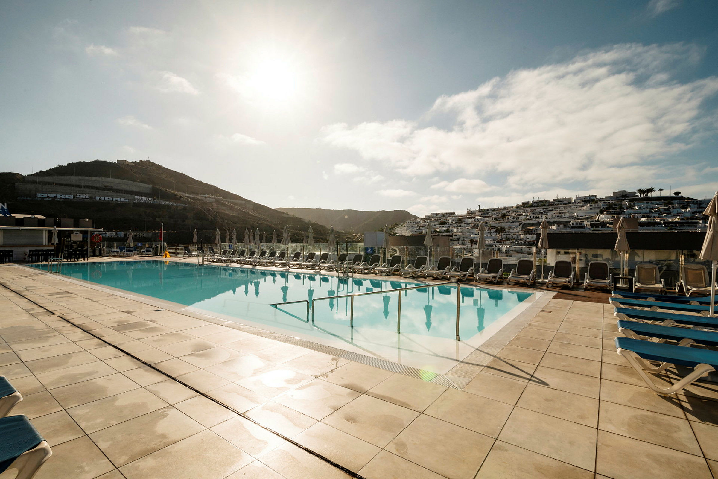 Udendørs pool med liggestole omkring og bjerge i baggrunden. (AI-genereret tekst)