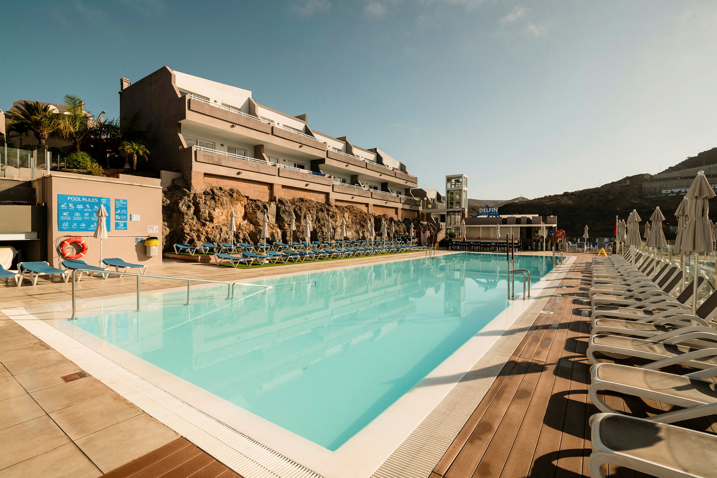 Udendørs swimmingpool med liggestole og hotelbygning i baggrunden. (AI-genereret tekst)