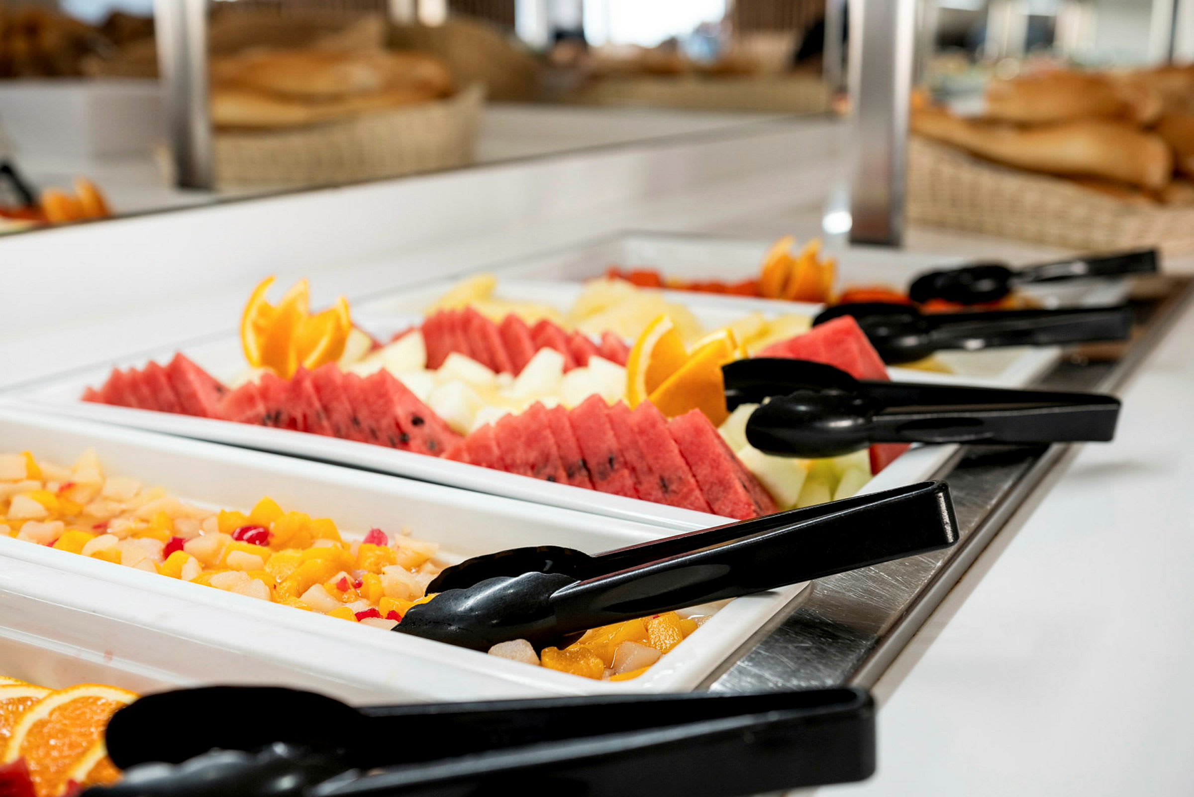 Buffetbord med frisk frugt og salat, inklusive meloner, appelsinskiver og tænger. (AI-genereret tekst)