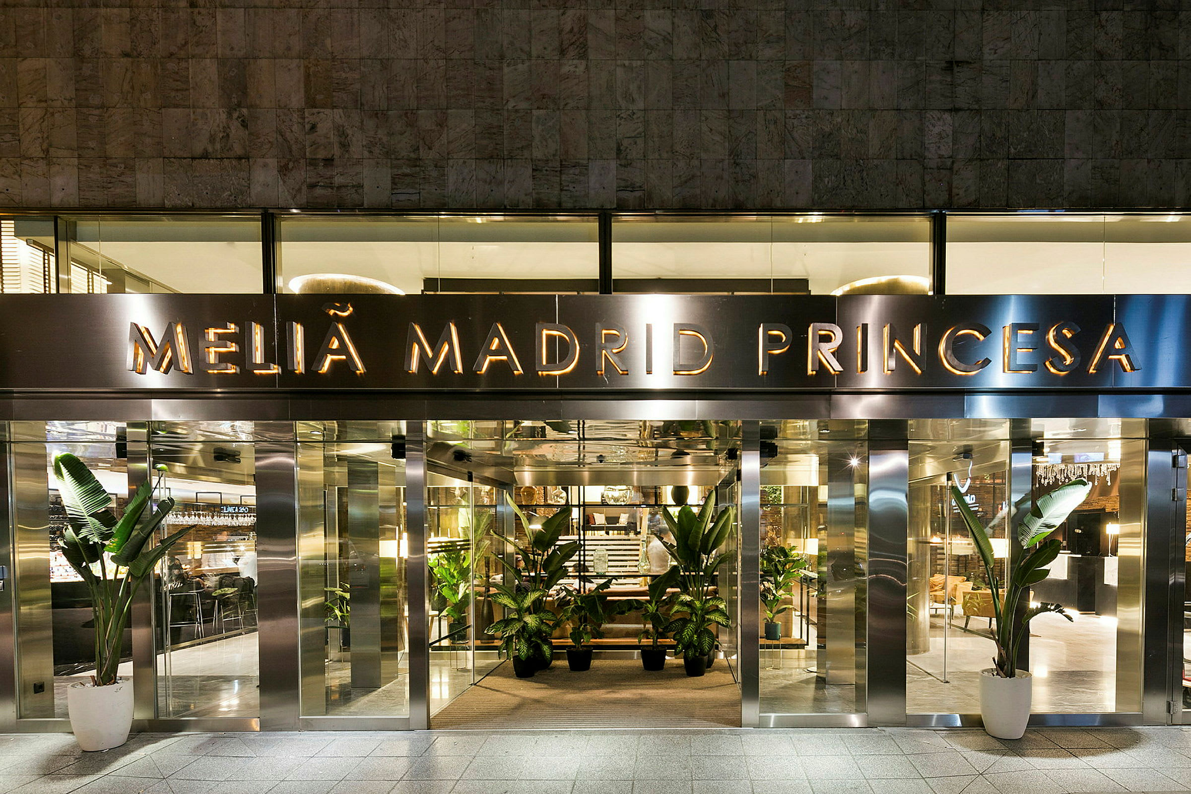 Indgangen til hotellet Melia Madrid Princesa med glasdøre og dekorative planter ved indgangen. (AI-genereret tekst)