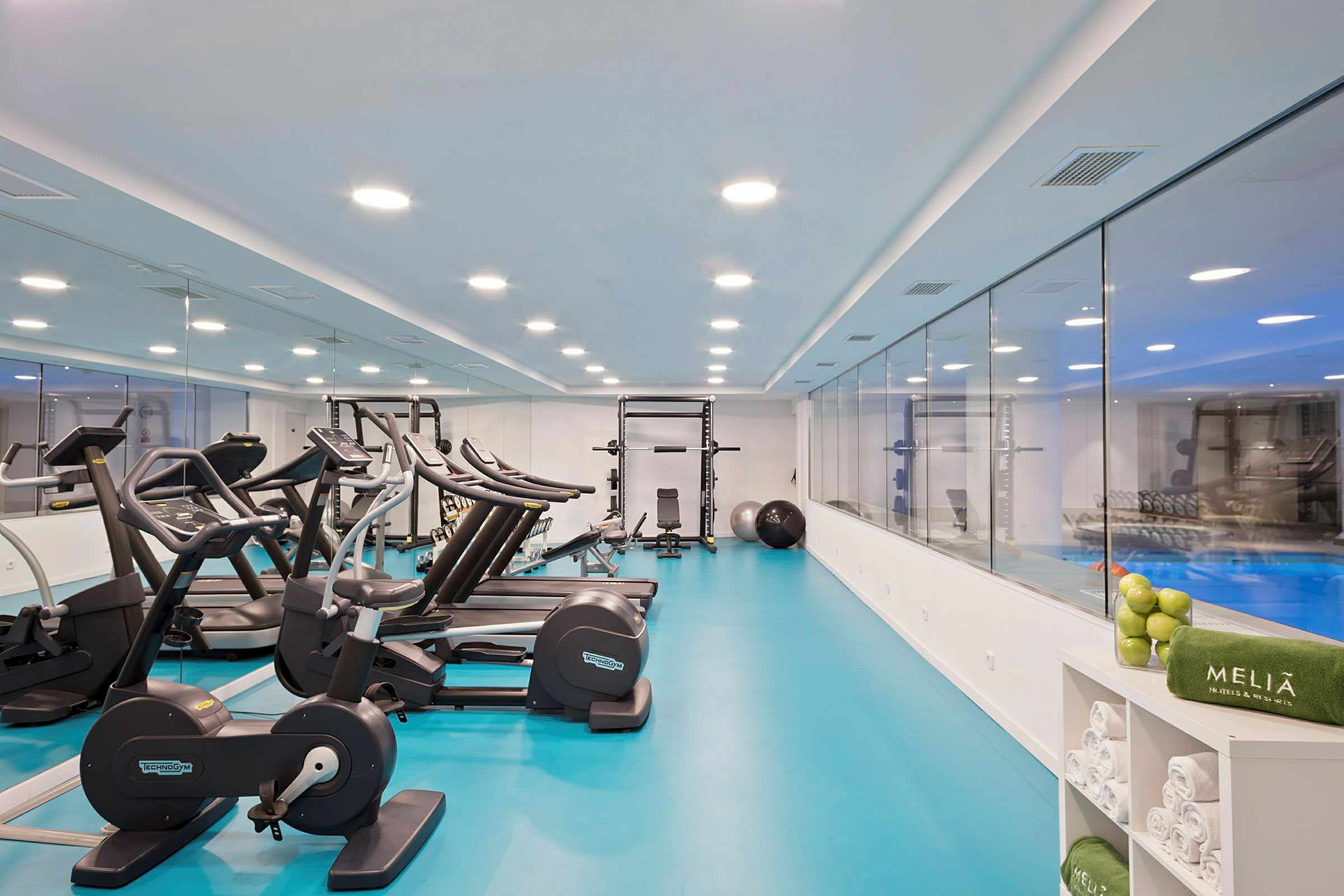 Et moderne fitnesscenter med træningsmaskiner, herunder løbebånd og motionscykler, samt en spejlvæg og en glasvæg der vender mod en pool. (AI-genereret tekst)