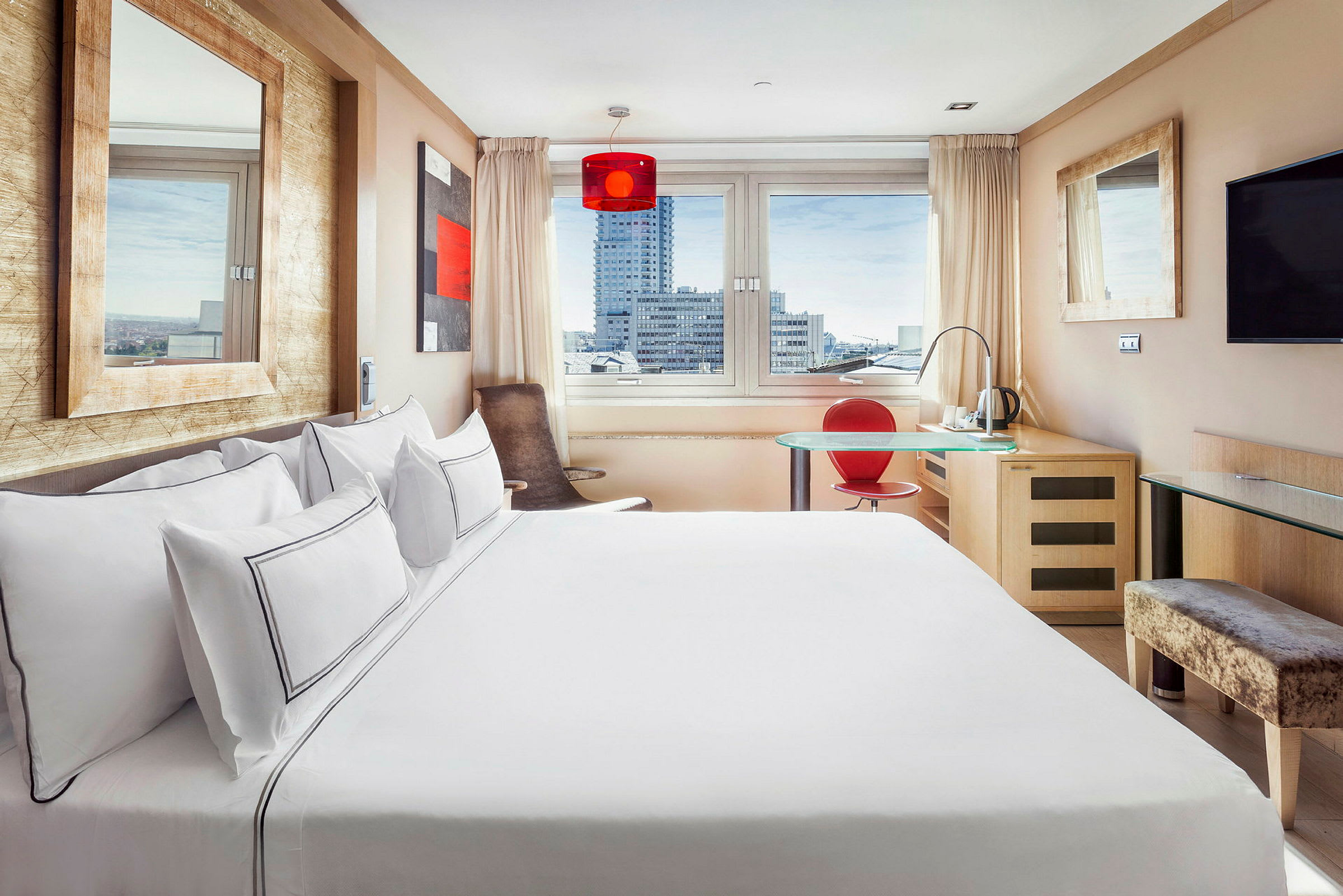 Et lyst og moderne hotelværelse med stor seng, vindue med udsigt over byens bygninger, et skrivebord med en rød stol og et vægmonteret TV. (AI-genereret tekst)