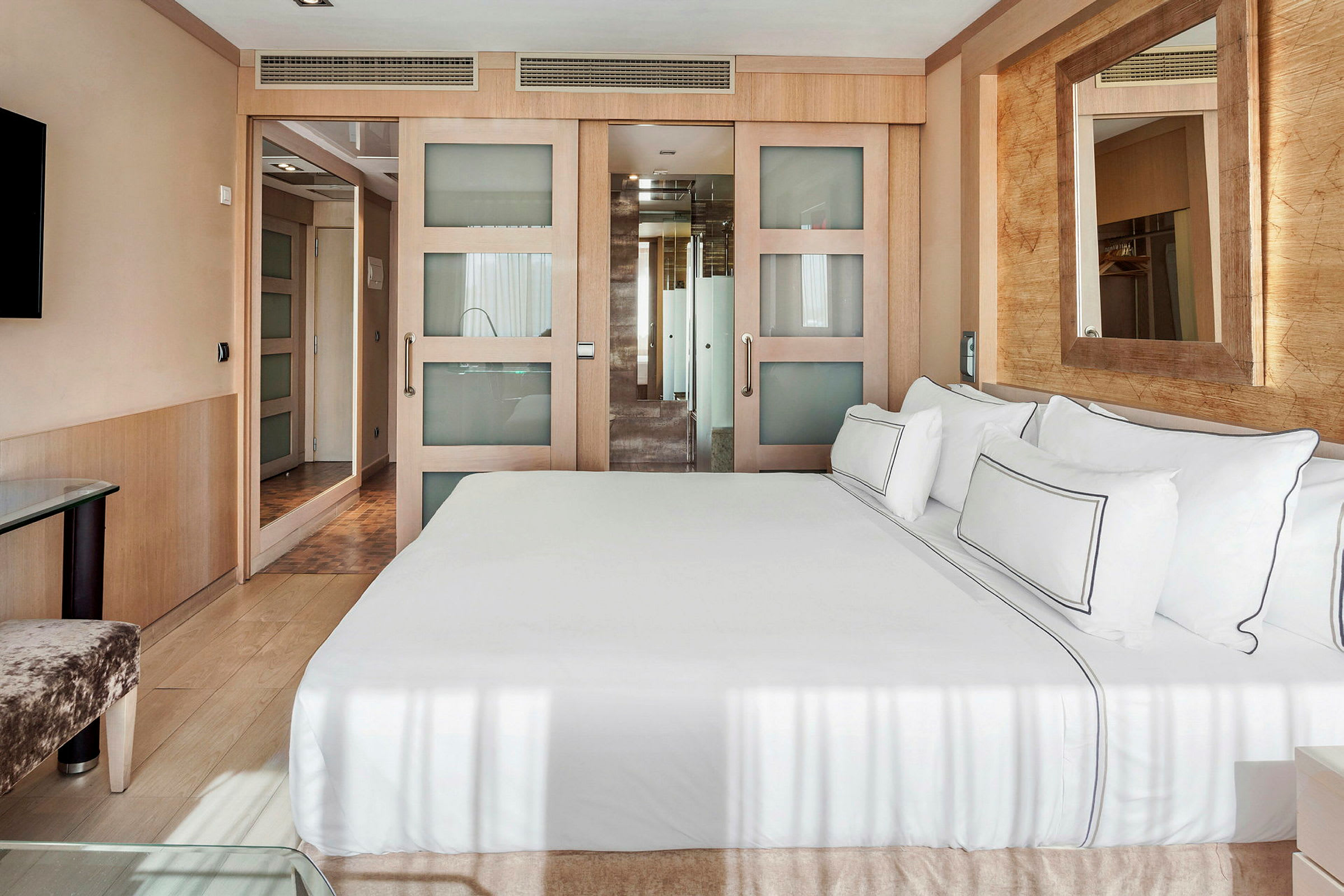 Et moderne hotelværelse med en stor seng med hvidt sengetøj, et spejl på væggen og en skydedør, der fører til et badeværelse. (AI-genereret tekst)