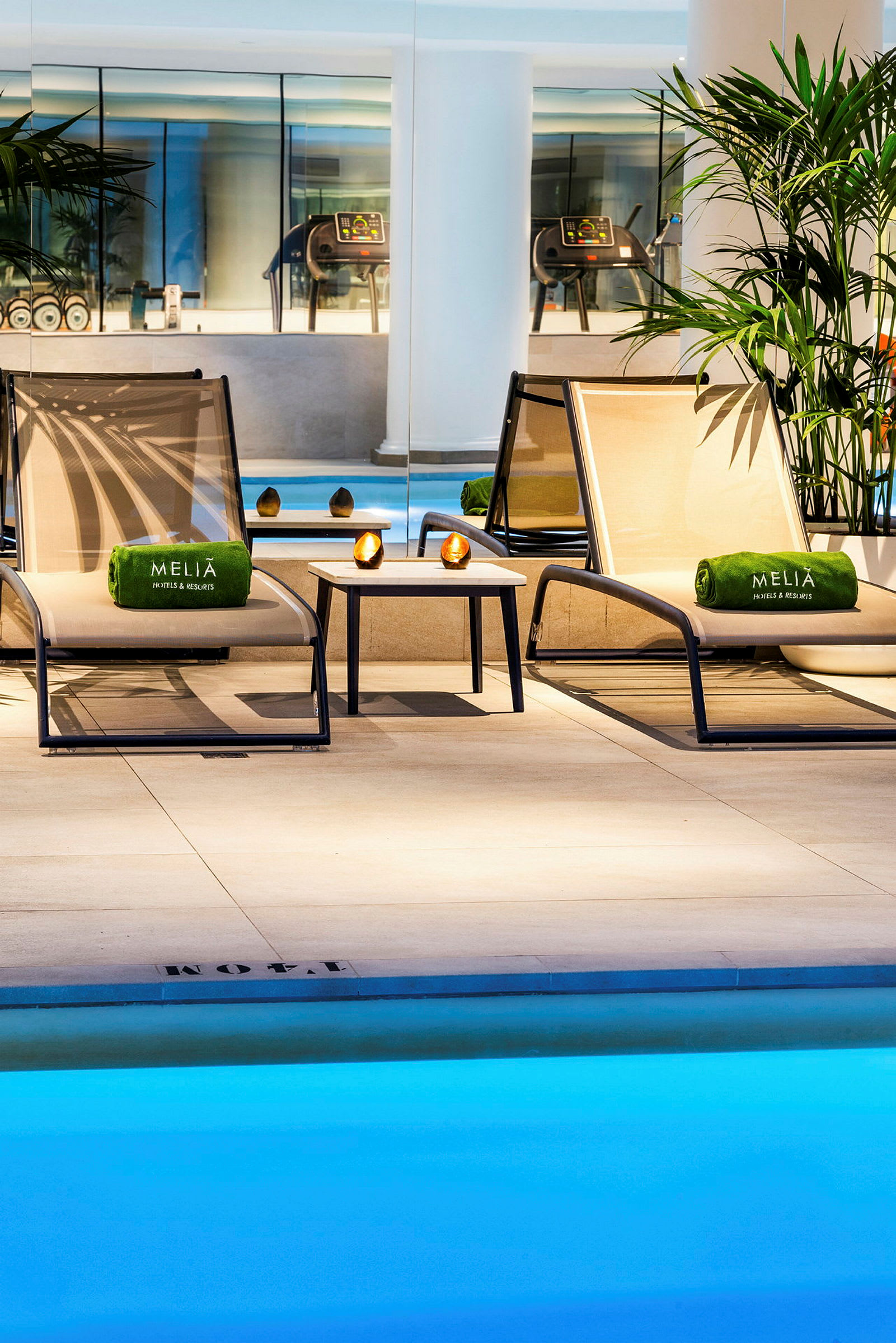 To solvogne med grønne håndklæder og et lille bord står ved en indendørs pool i et elegant spaområde. (AI-genereret tekst)