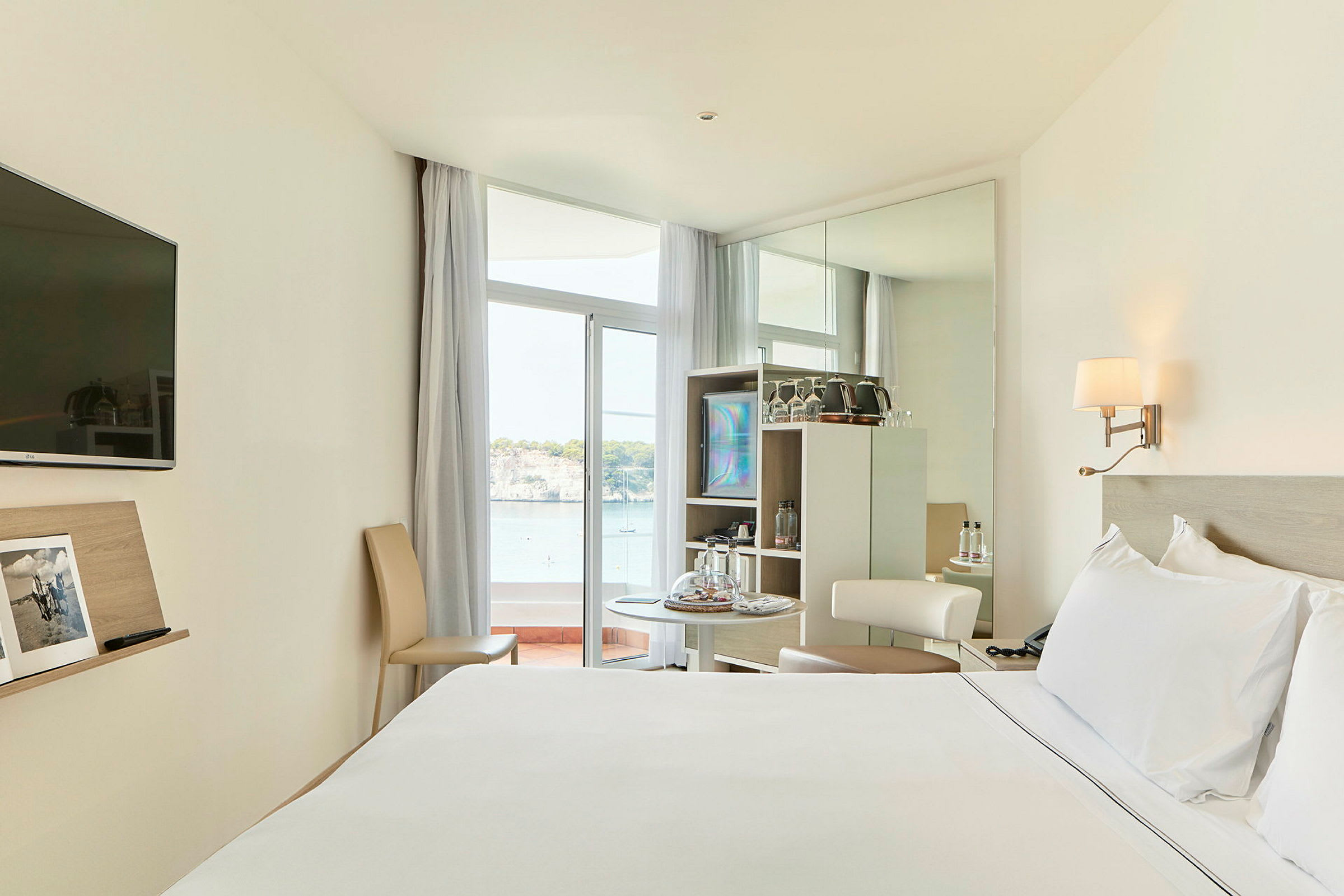 Et hotelværelse med en stor seng, et lille bord med stole og udsigt over havet gennem en glasdør. (AI-genereret tekst)