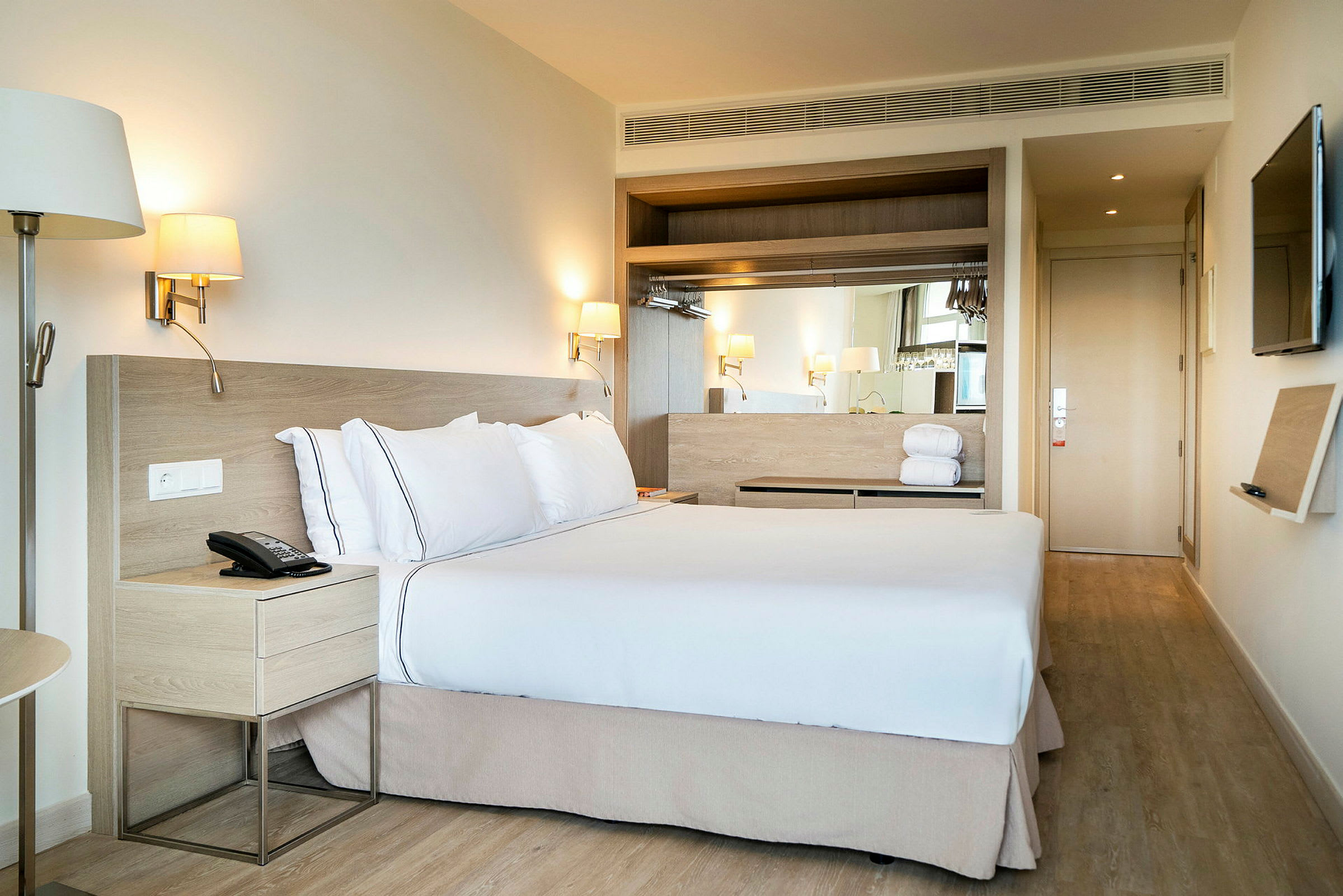 Et hotelværelse med en stor seng redt op med hvide lagner og puder. Lamper på væggen over siderne af sengen samt et natbord ved siden af. (AI-genereret tekst)
