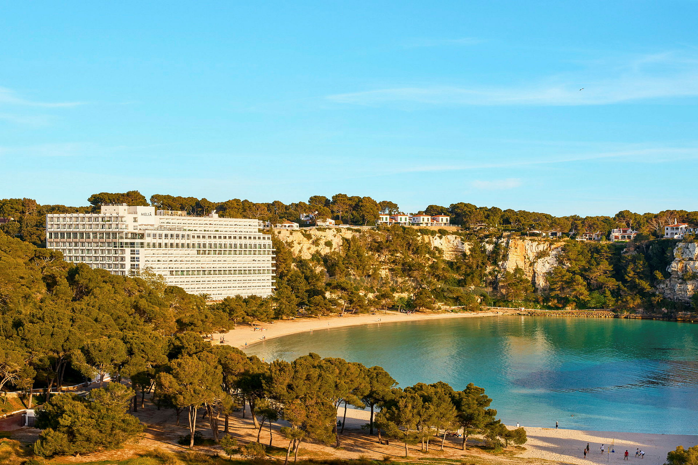 Hotelbygning ved en klippekyst med en sandstrand og turkisblåt vand. (AI-genereret tekst)