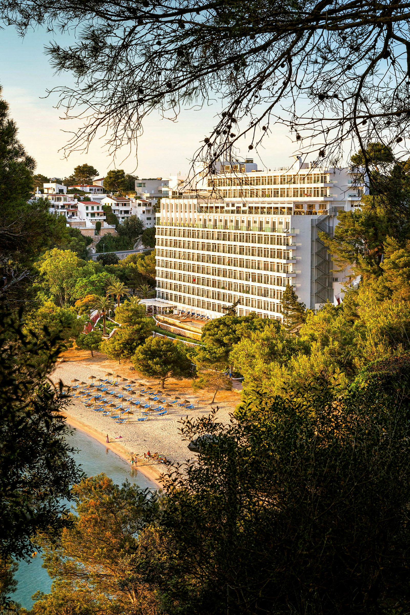 Billede af et hotel beliggende tæt på en strand med liggestole og parasoller, omgivet af frodige træer. (AI-genereret tekst)