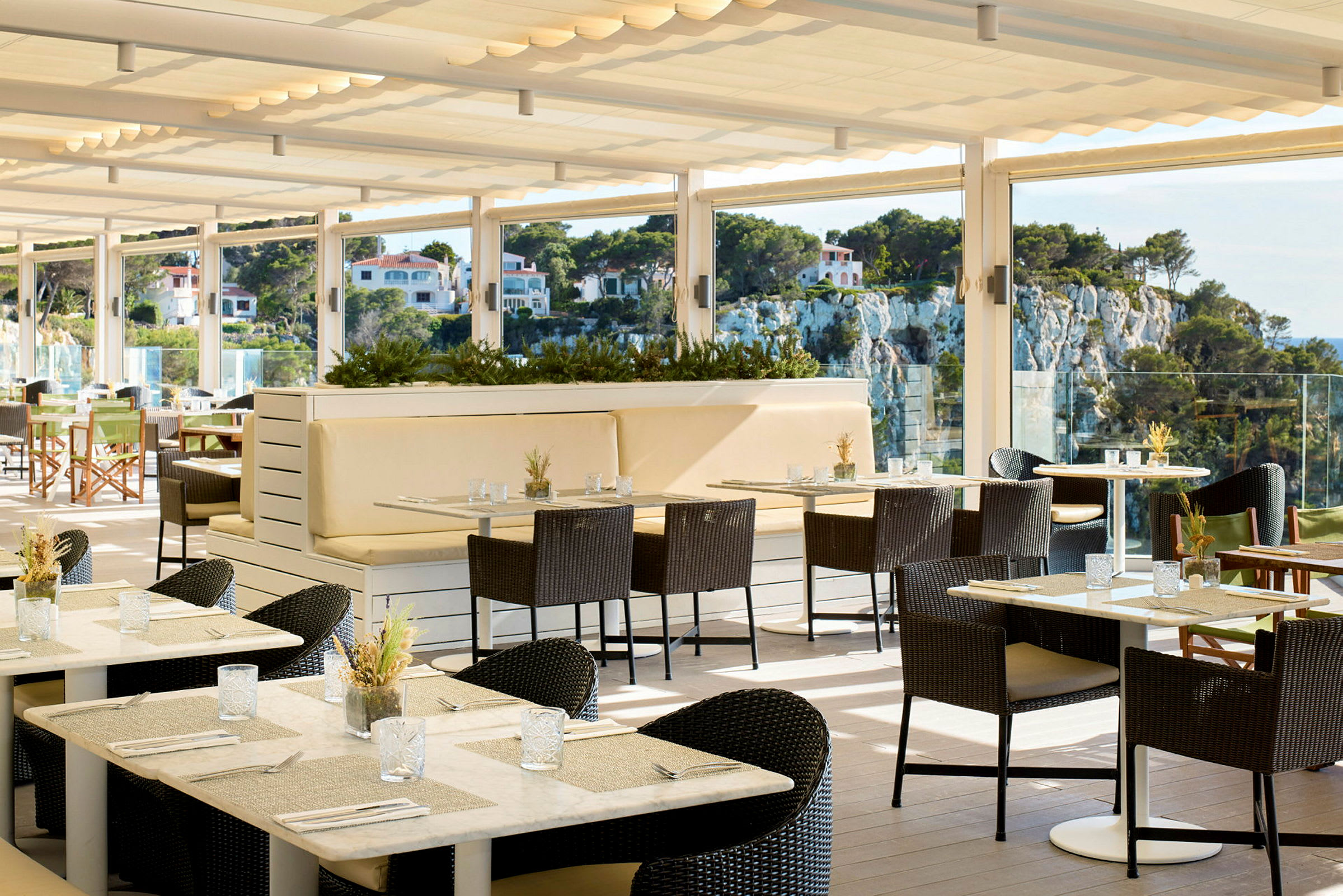 En solrig restaurantterrasse med borde og stole samt udsigt over et frodigt område med huse på en klippe. (AI-genereret tekst)