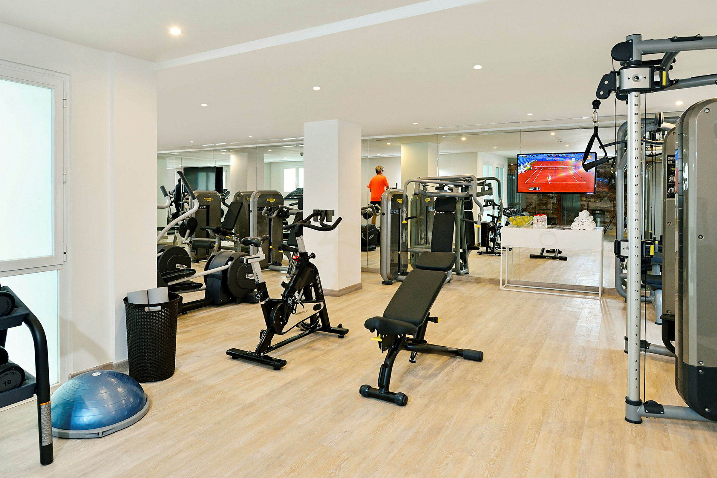 Et fitnesscenter med træningsudstyr såsom cykler, bænke og maskiner samt en TV på væggen. (AI-genereret tekst)