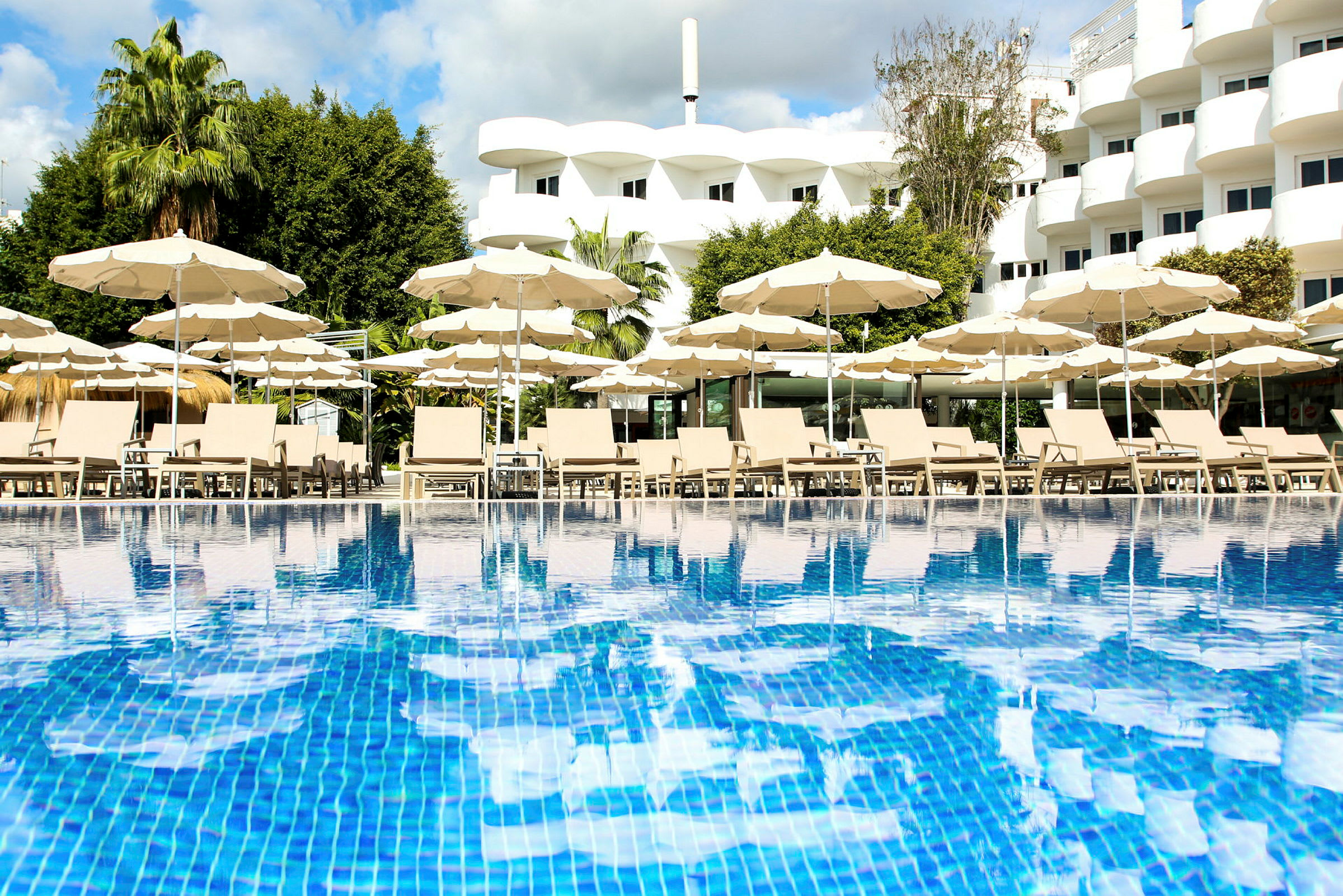 En pool med liggestole og parasoller foran et hotel. (AI-genereret tekst)