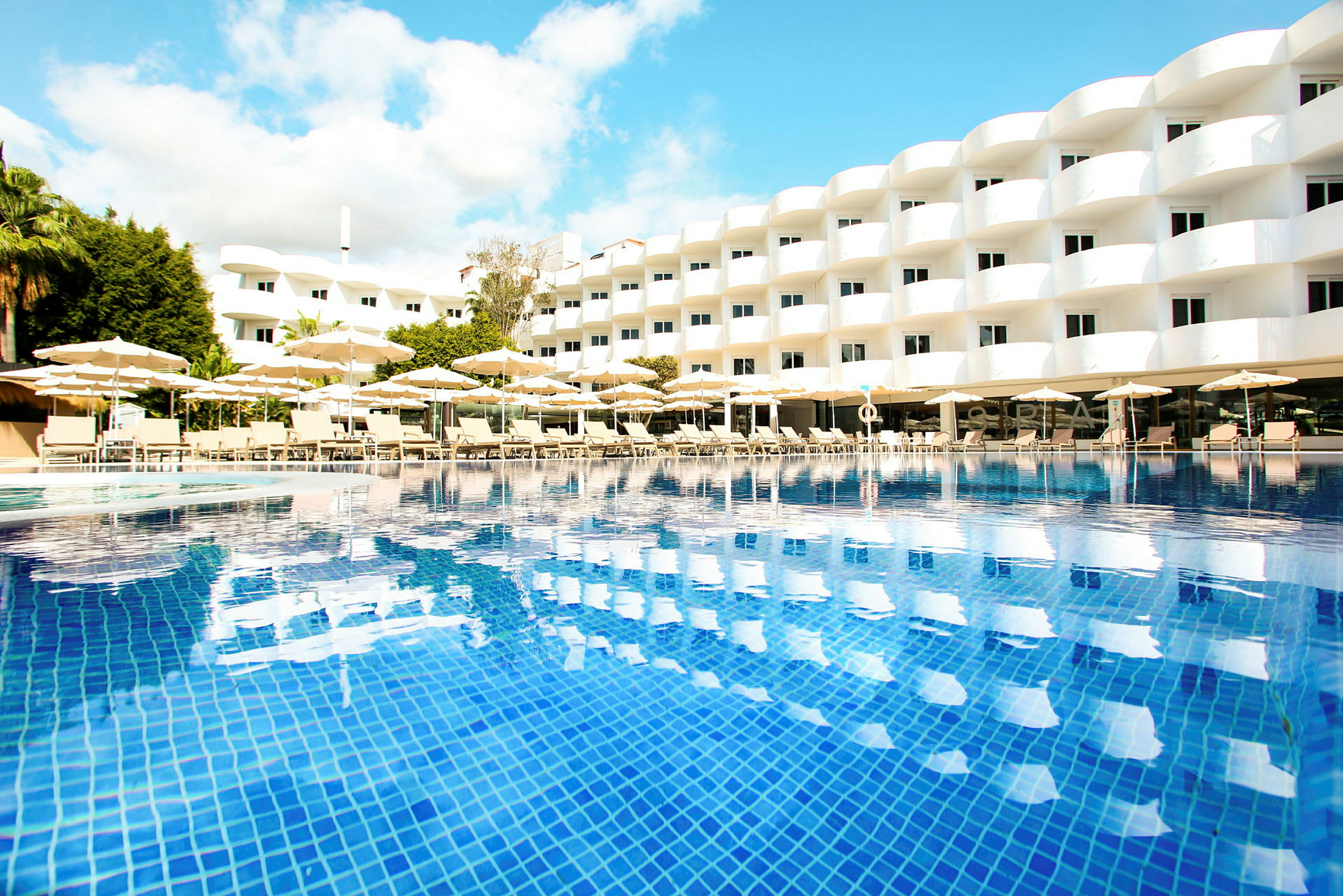 Et moderne hotel med strålende hvide facader, liggestole og parasoller ved en klarblå pool. (AI-genereret tekst)