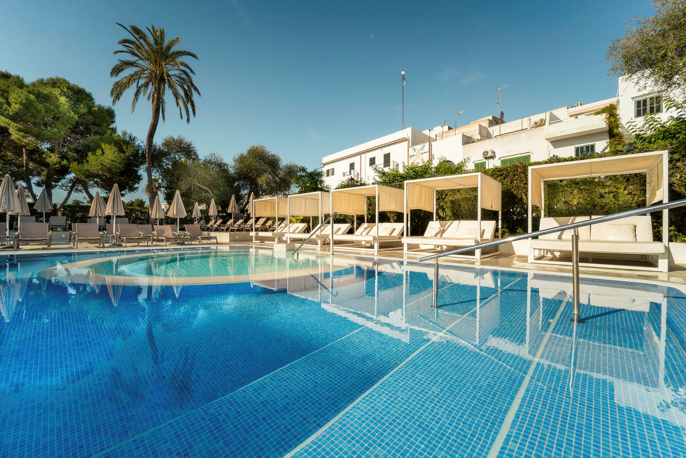 Billede af en pool med turkis vand samt hvide solsenge under parasoller og cabanas. Der er omkringliggende grønt og en skygge af bygninger i baggrunden. (AI-genereret tekst)