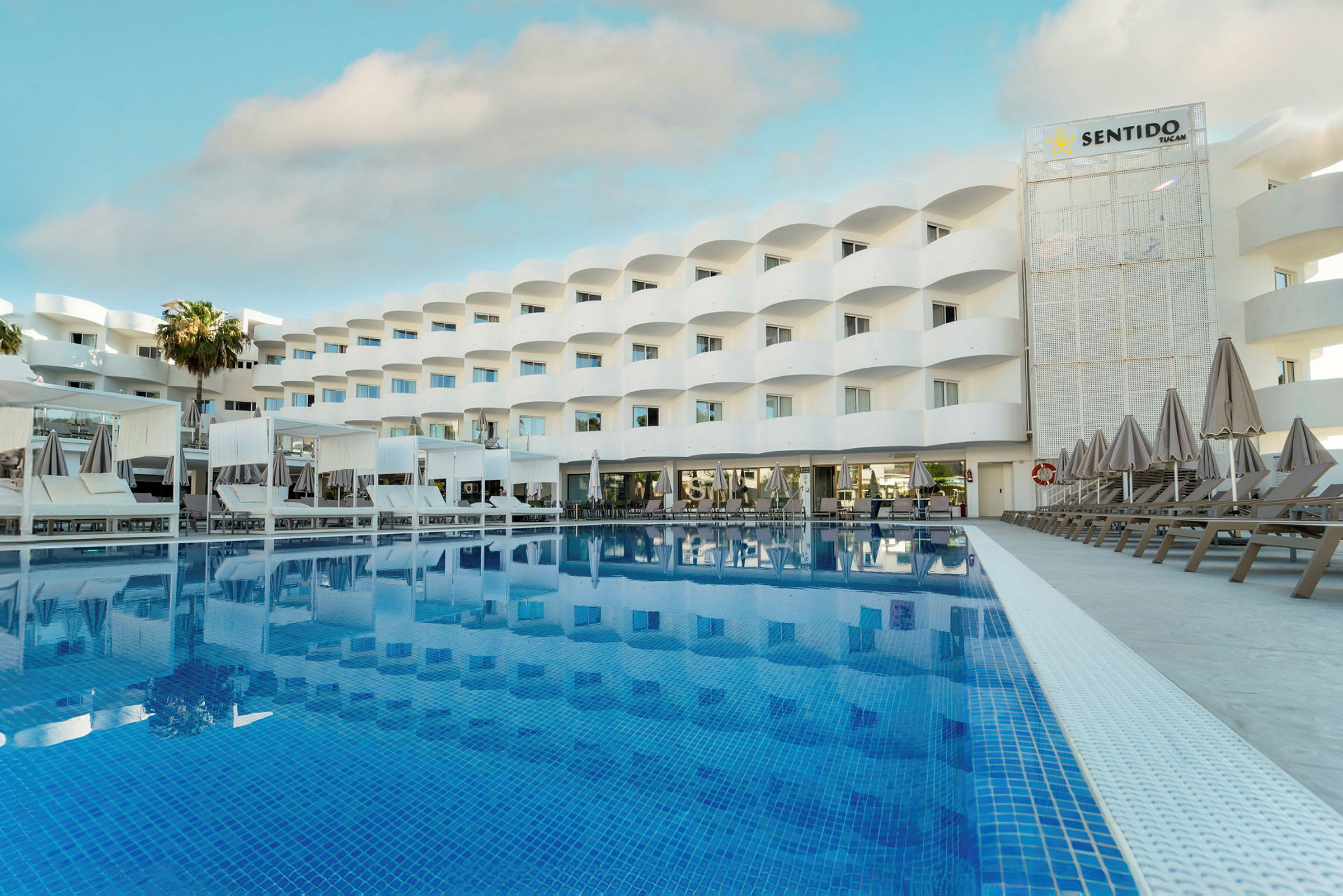 Billede af et hotel med en stor pool foran. Bygningen er hvid med altaner og liggestole ved poolområdet. (AI-genereret tekst)