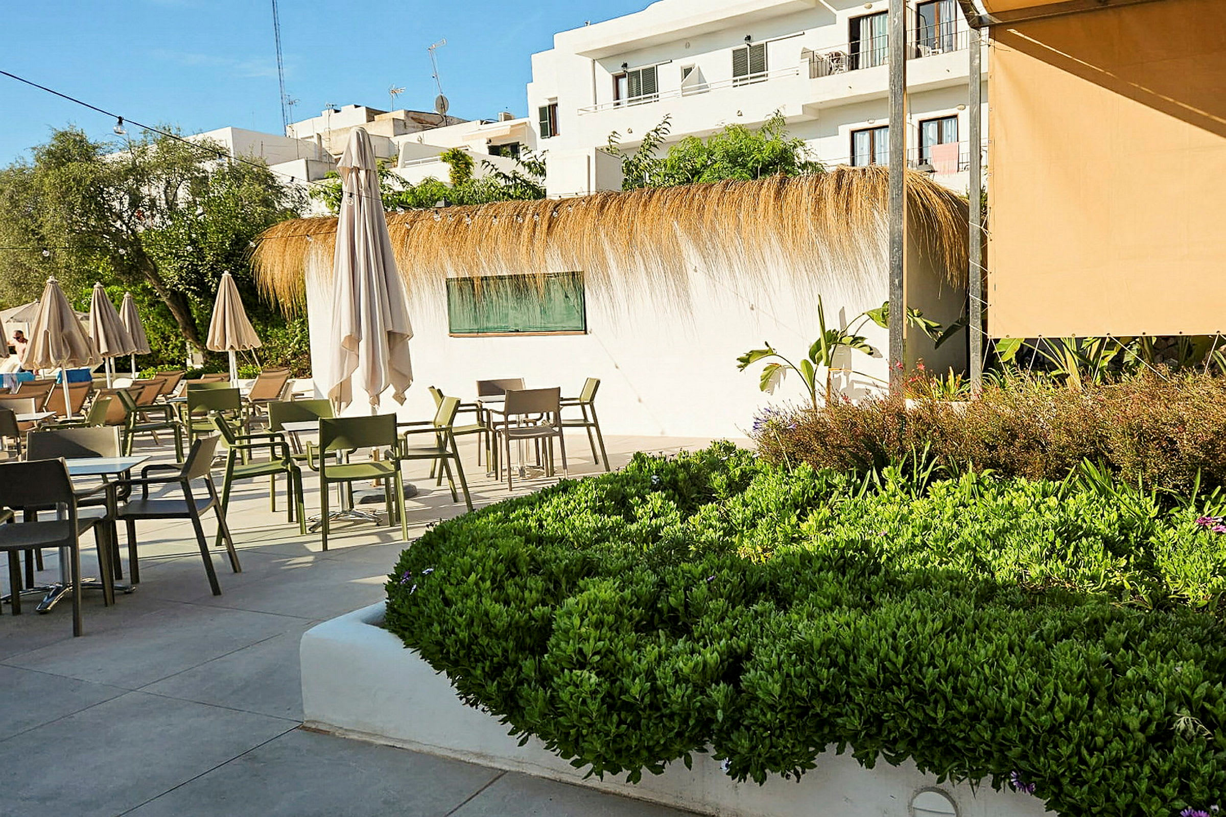 En udendørs terrasse med borde, stole og parasoller samt beplantning og hvide bygninger i baggrunden. (AI-genereret tekst)